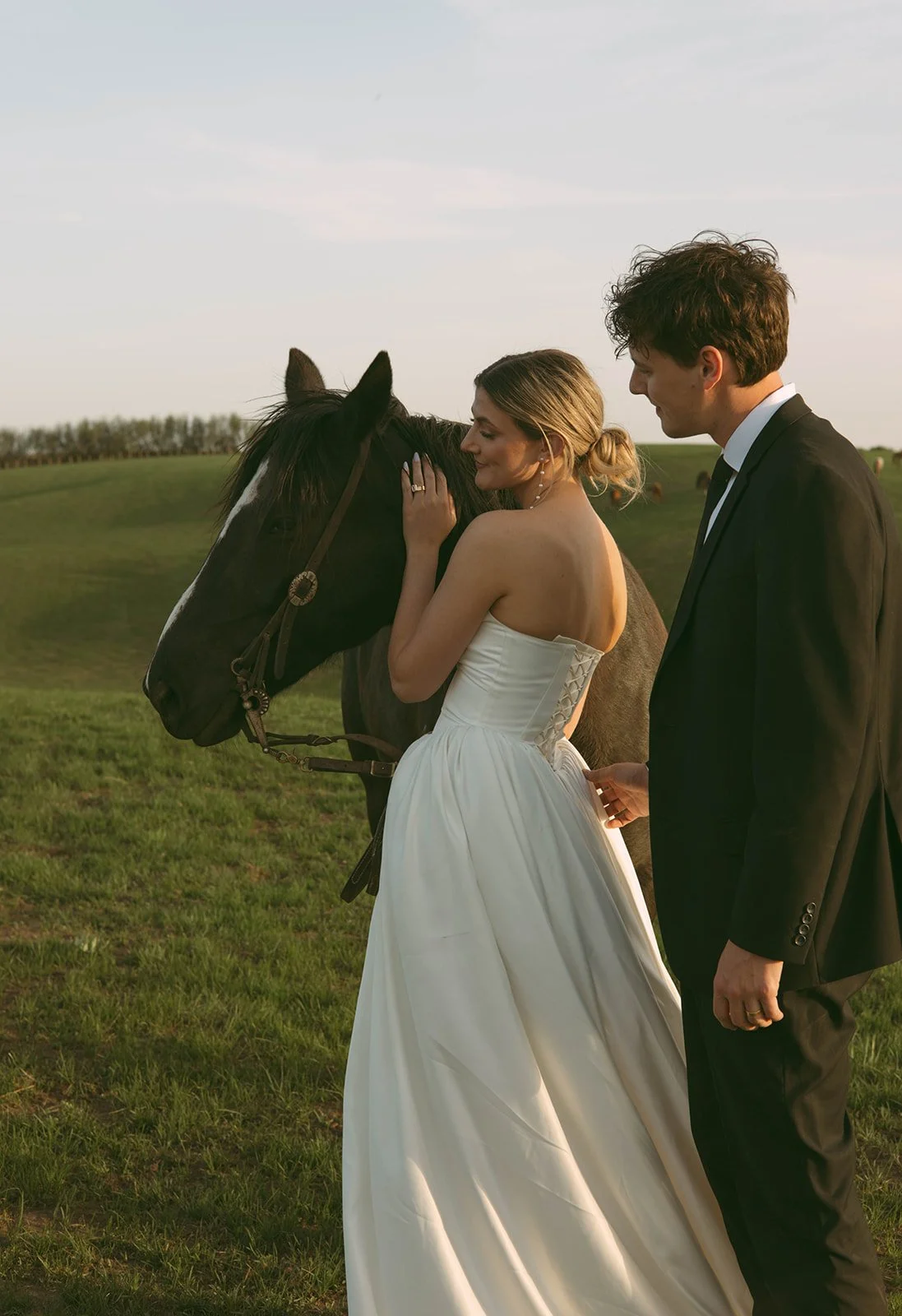 A bride in a white wedding dress gently touching a horse's face, with a groom standing beside her, in an open grassy field at sunset.