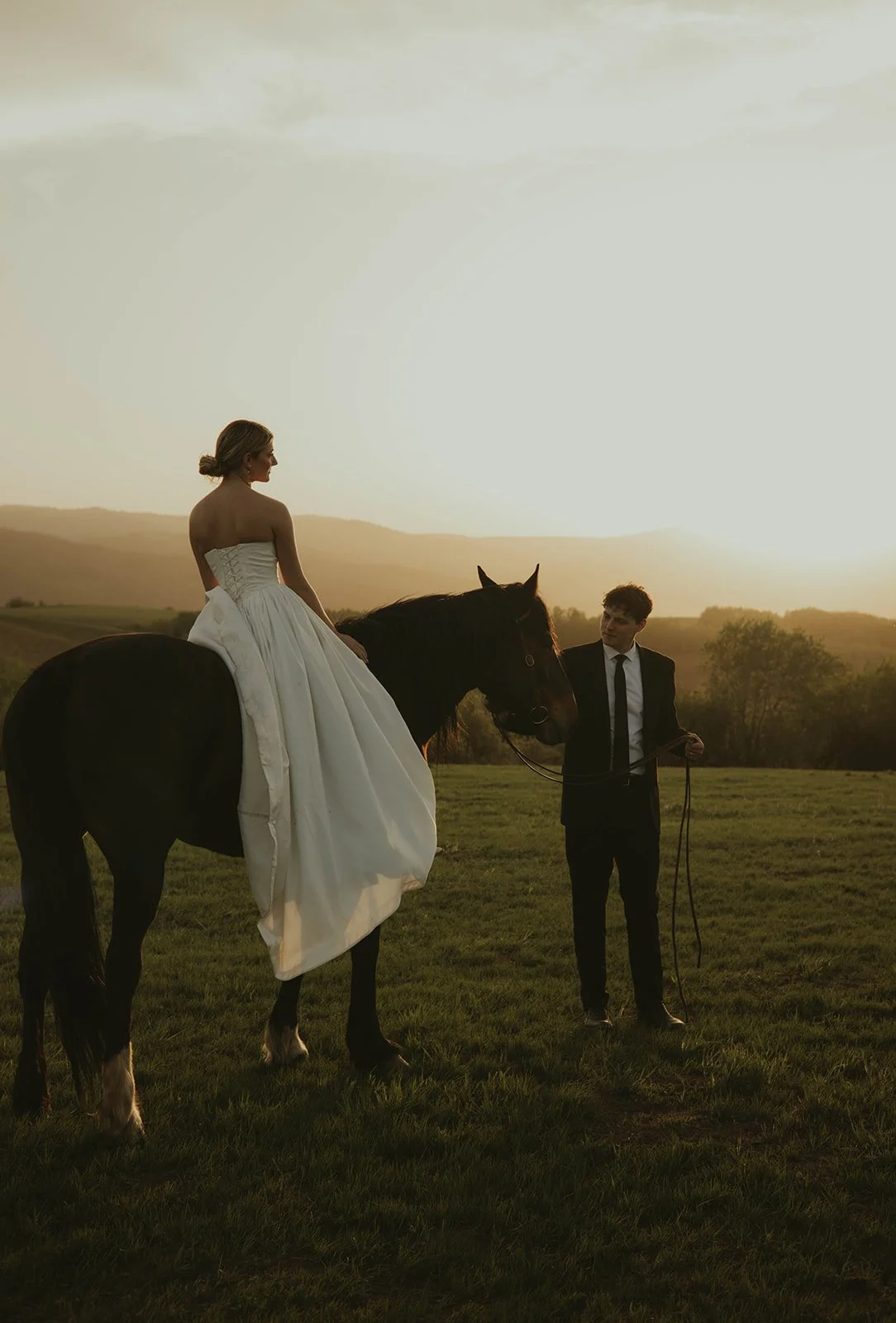A woman in a white wedding dress sitting on a black horse, with a man in a suit holding the horse's reins during sunset in a field.