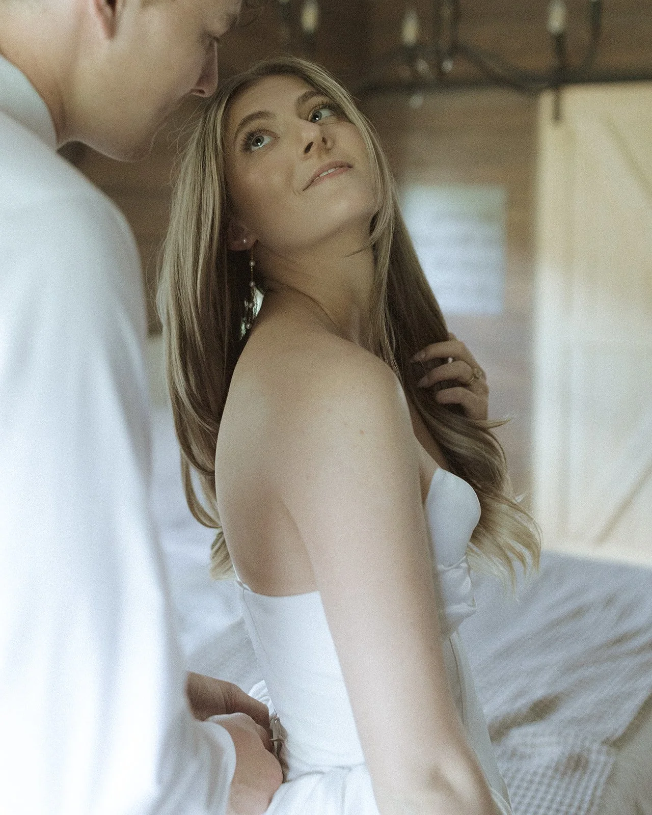 A young woman with long blonde hair sitting on a bed, looking over her shoulder at a man who is partially visible.