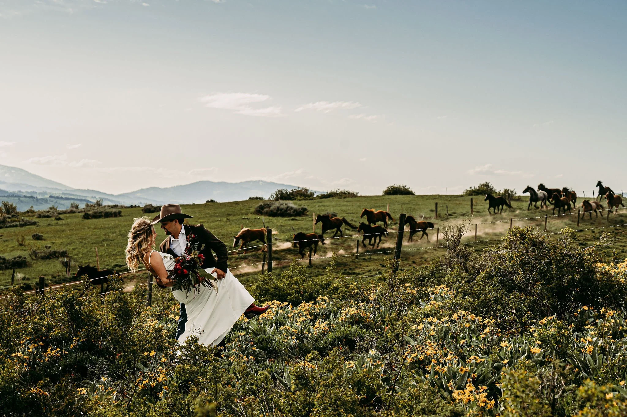 A newlywed couple in wedding attire, with the groom wearing a cowboy hat, sharing a dance in a field of yellow flowers, with a herd of horses running in the background on a partly cloudy day.