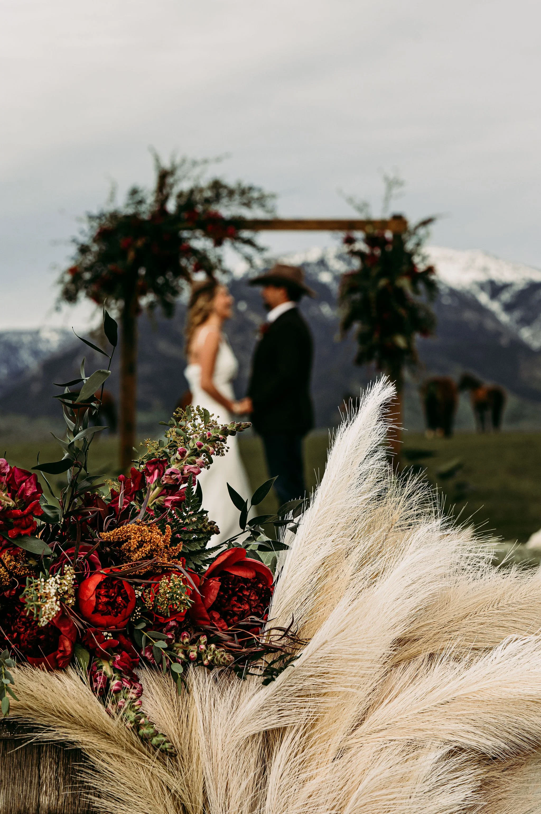 A close-up of a wedding bouquet with red flowers and pampas grass in the foreground, and a bride and groom holding hands in the background, with mountains and horses behind them.
