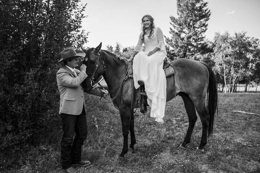 A woman in a white dress is sitting on a horse, smiling, while a man in a cowboy hat and jacket stands next to the horse, holding its bridle.