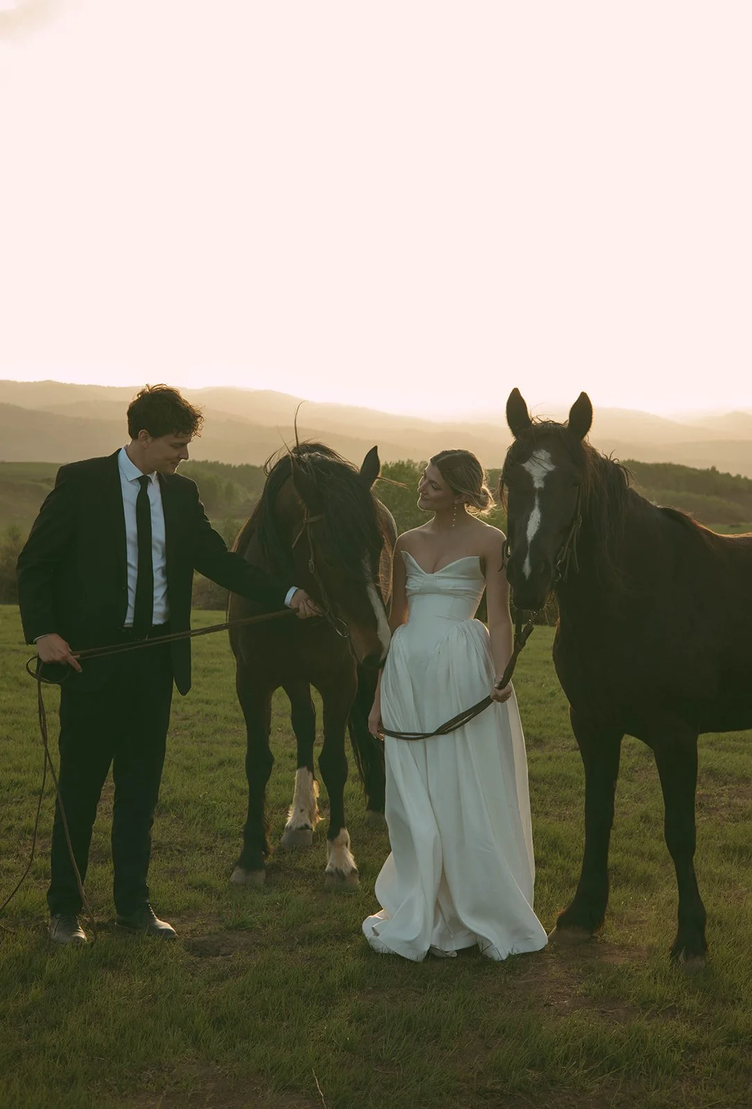 A man in a black suit and tie and a woman in a white dress standing outdoors on a grassy field with two horses during sunset.