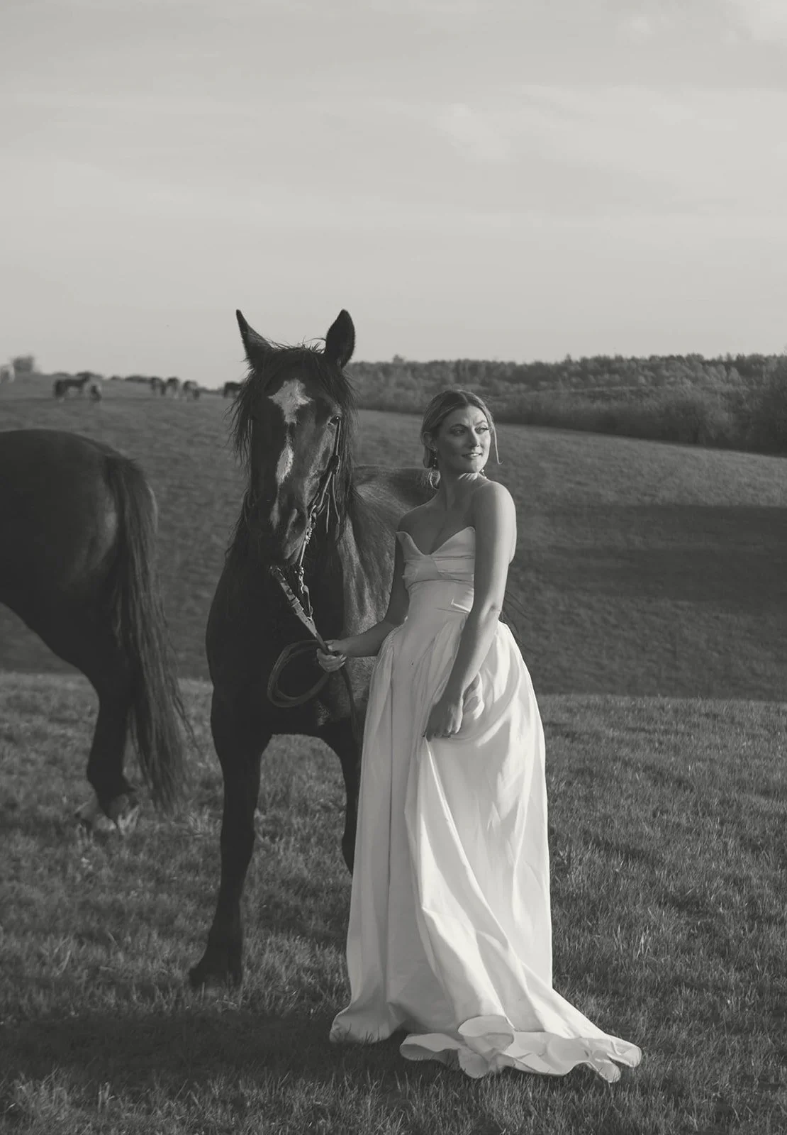 A woman in a white strapless dress holding a horse's lead in a grassy field, with other horses grazing in the background, in black and white.