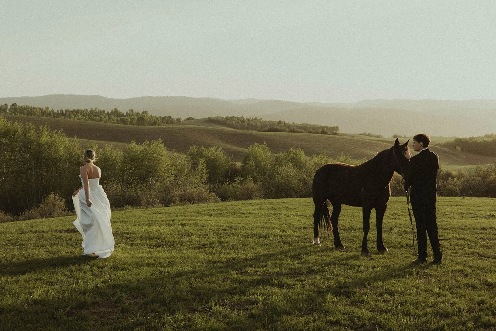 A woman in a white dress walking across a grassy field towards a man in a black suit who is standing next to a black horse in a scenic rural landscape at sunset.