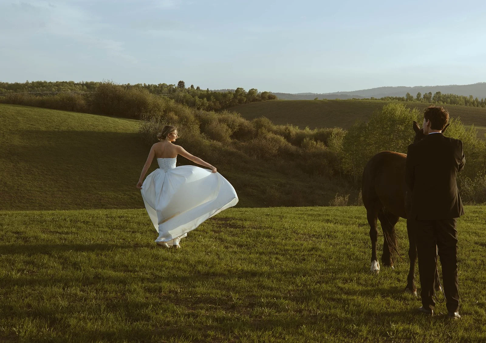 A woman in a wedding dress standing on a grassy field with a man in a suit holding the reins of a horse during sunset, with rolling hills and trees in the background.