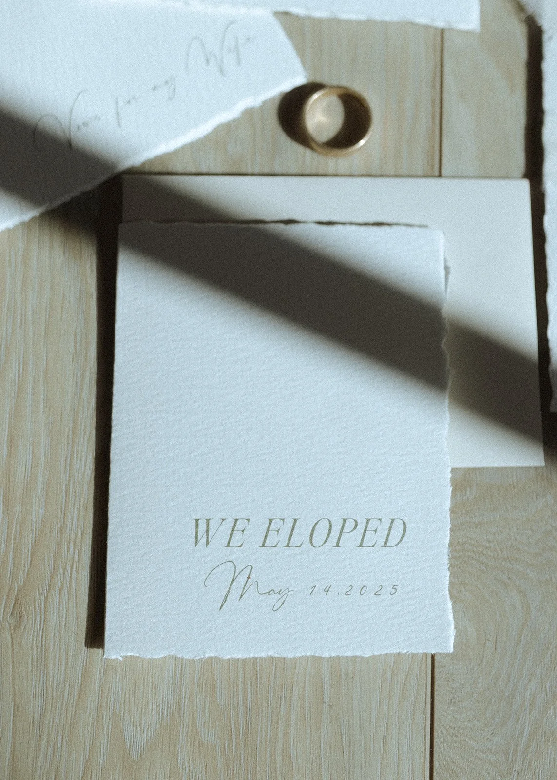 A torn piece of white paper on a light wood surface with the words "WE ELOPED May 14, 2025" printed in elegant font.