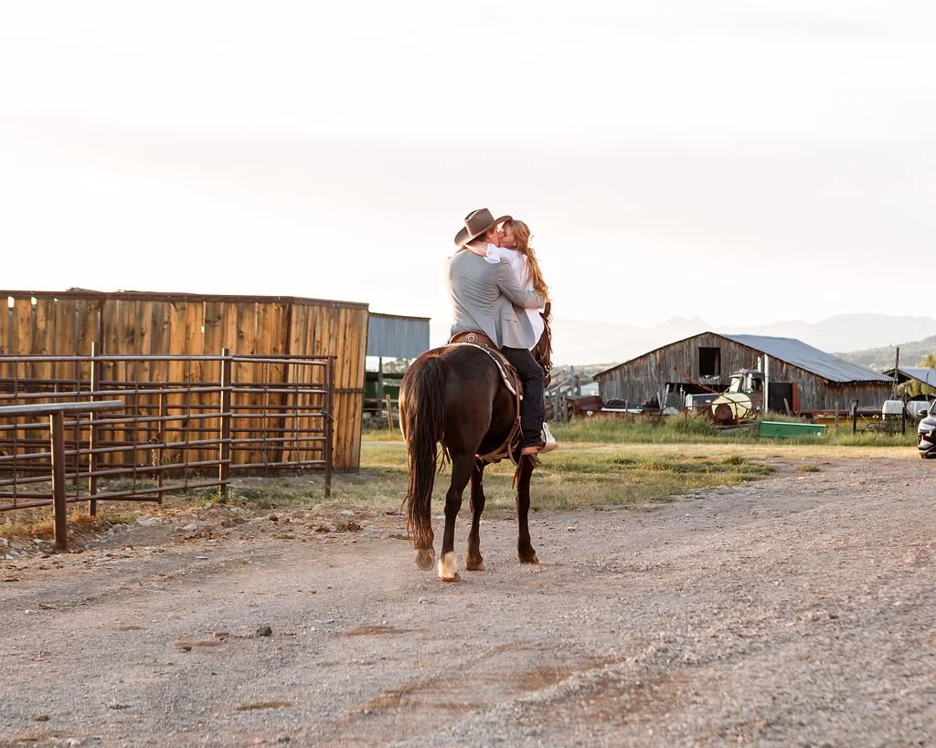 A couple on horseback embracing near a farm with barns and equipment, during sunset.