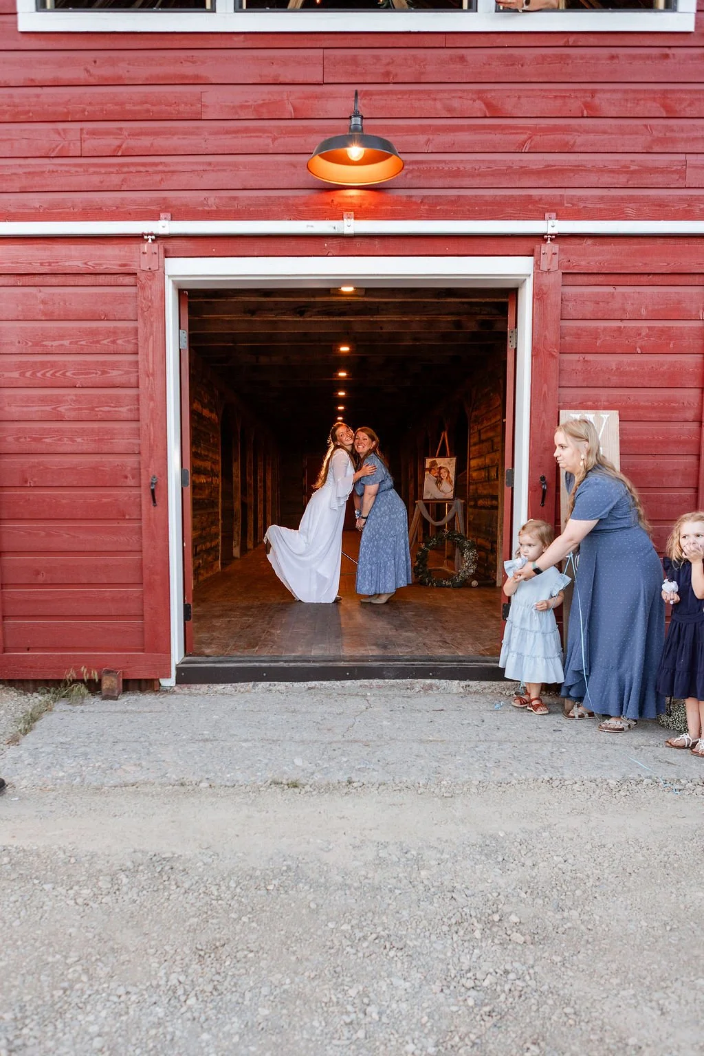Two women hugging inside a rustic barn with red exterior, with several children and a woman watching on the side.