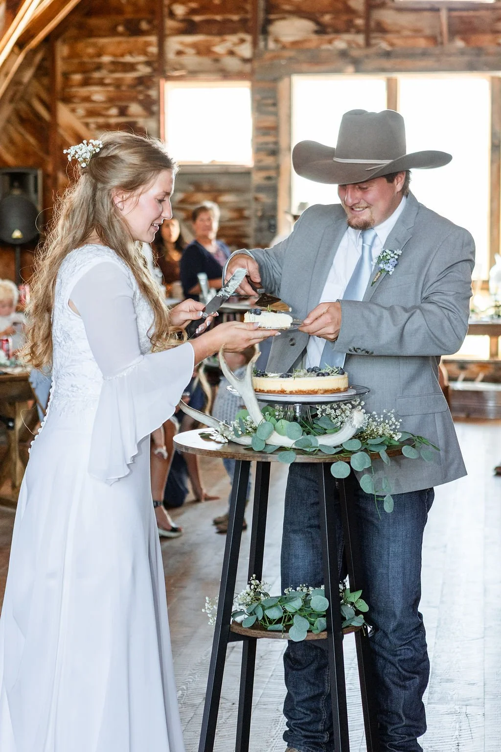 A bride and groom at a wedding celebration are cutting and serving wedding cake. The bride is wearing a white wedding dress, and the groom is dressed in a gray suit with a large cowboy hat, in a rustic barn setting with guests in the background.
