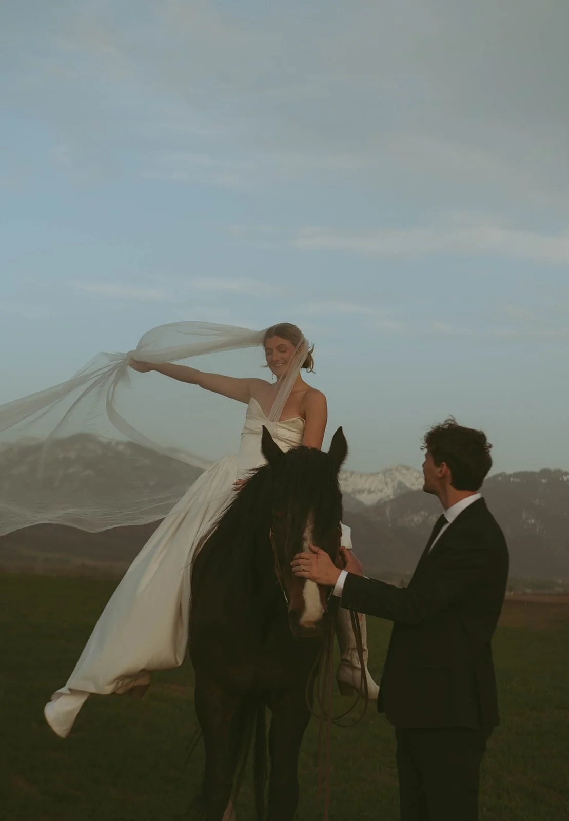 A woman in a wedding dress sitting on a black horse, holding a flowing white veil, with a man in a black suit standing nearby, holding the horse's reins, in an open field with mountains in the background.
