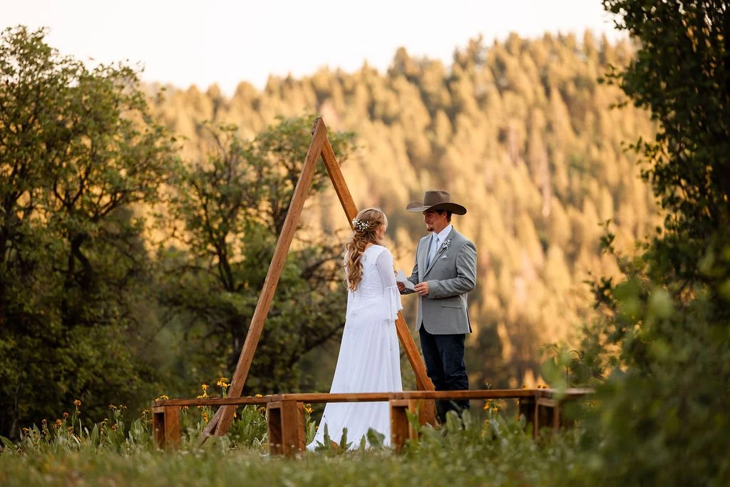A couple exchanging vows in an outdoor wedding ceremony, officiated by a man in a cowboy hat, surrounded by trees and mountains in the background.