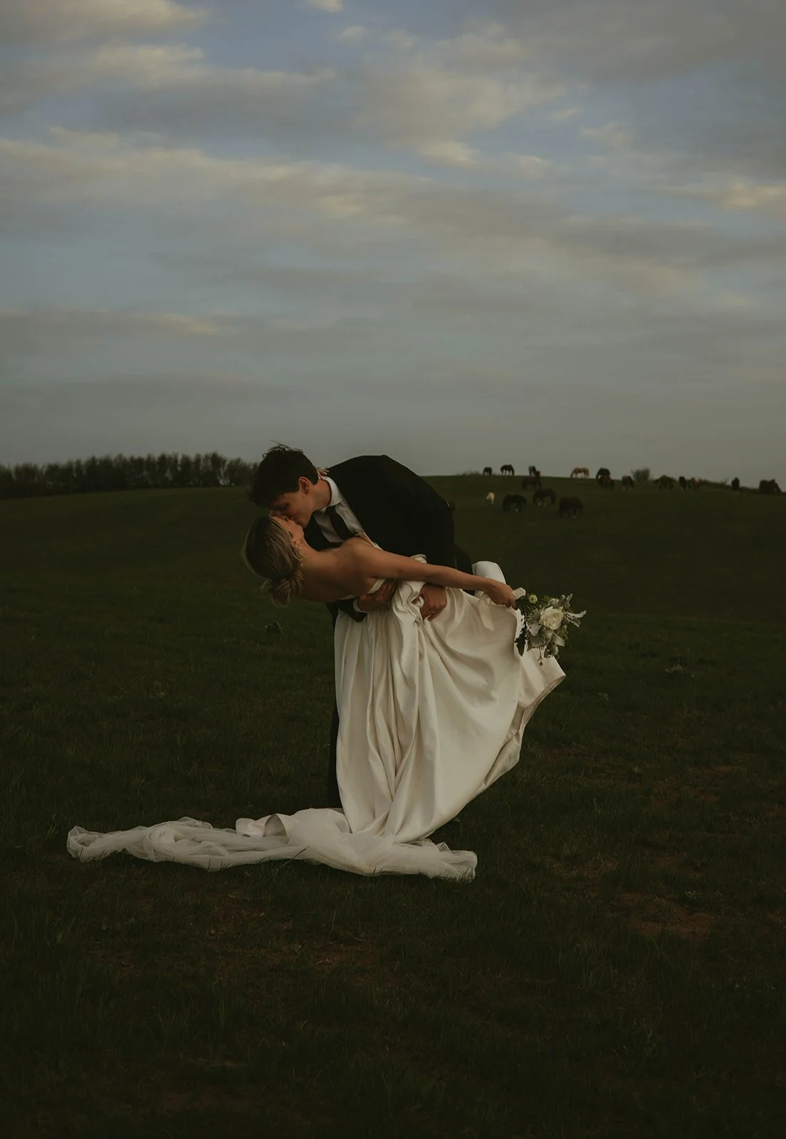 A groom holding and kissing a bride in a field with horses in the background during sunset or early evening.