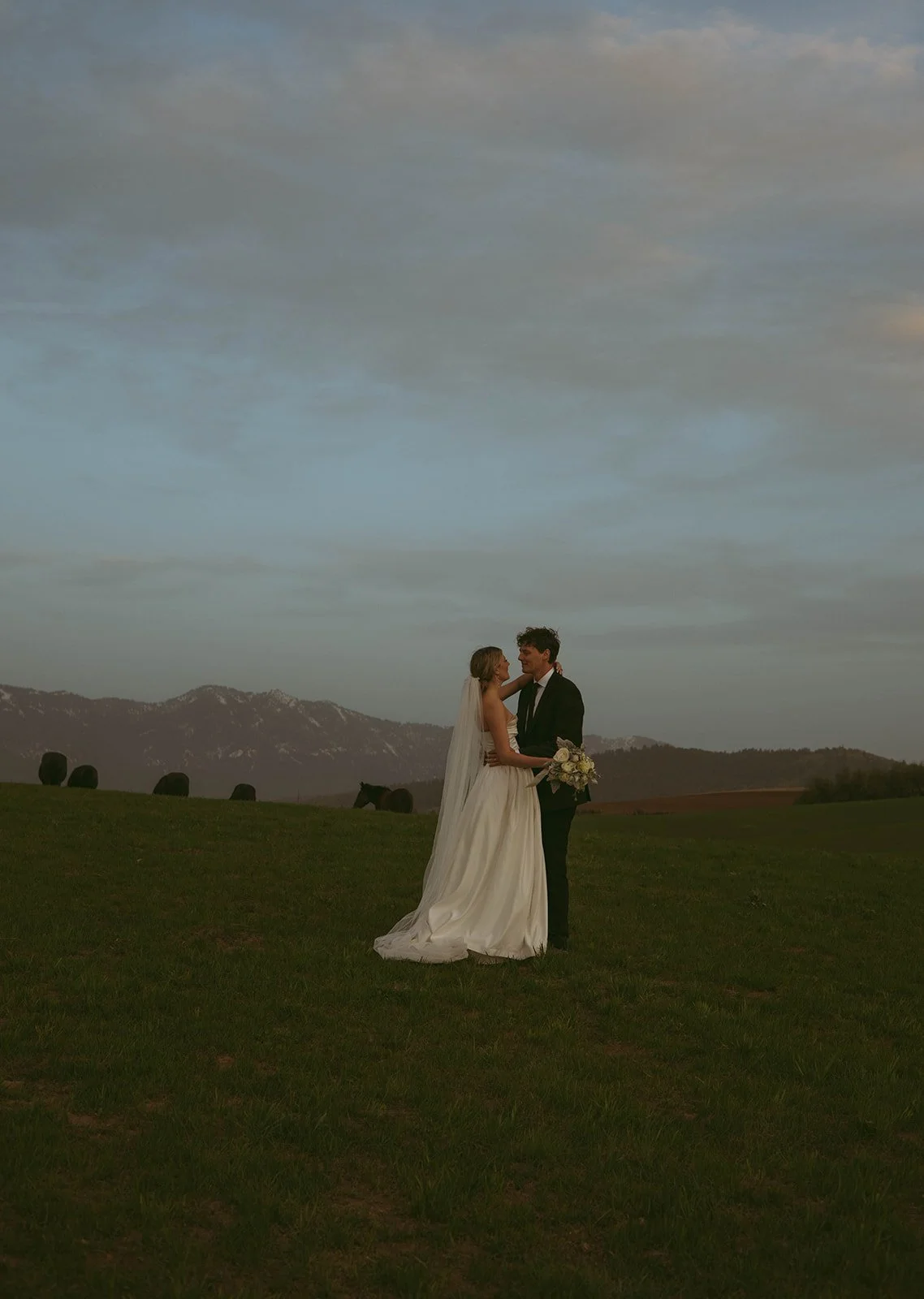 A bride and groom in wedding attire sharing a kiss outdoors with a mountain range in the background during sunset.