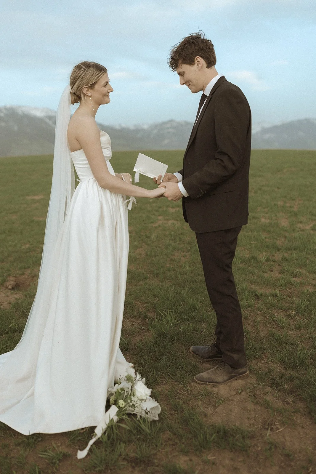 A bride and groom holding hands during their outdoor wedding ceremony, standing on a grassy field with mountains in the background.