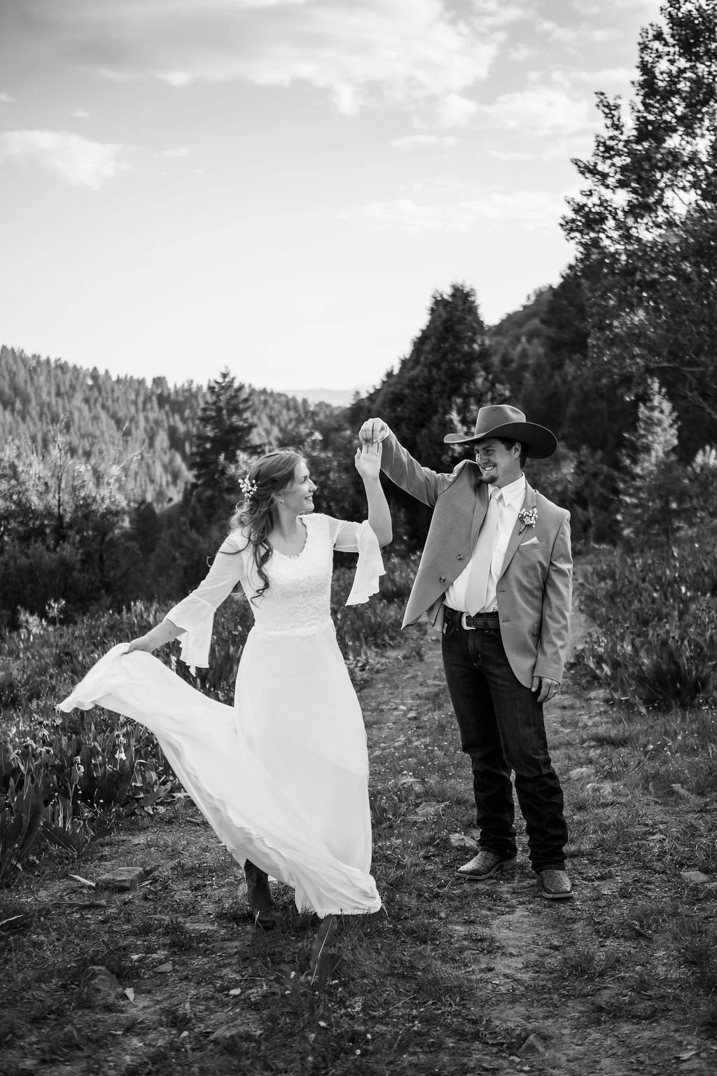 A black and white photo of a bride and groom dancing outdoors on a dirt path surrounded by trees and mountains, with the groom wearing a cowboy hat and the bride in a white dress.