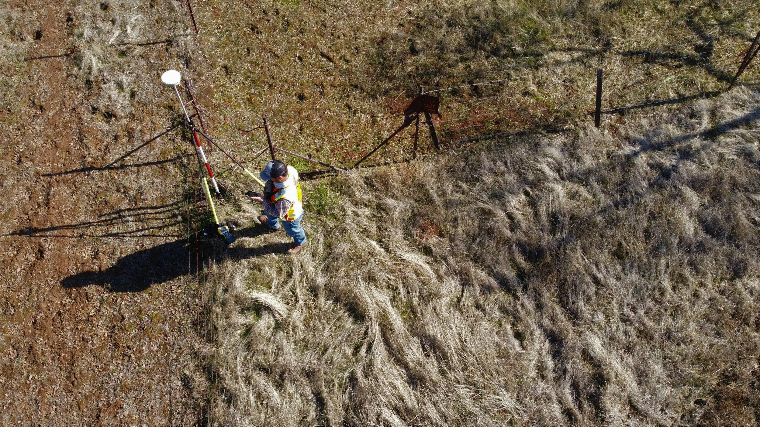 A person standing near a survey instrument and equipment in a grassy, dry field, viewed from above.
