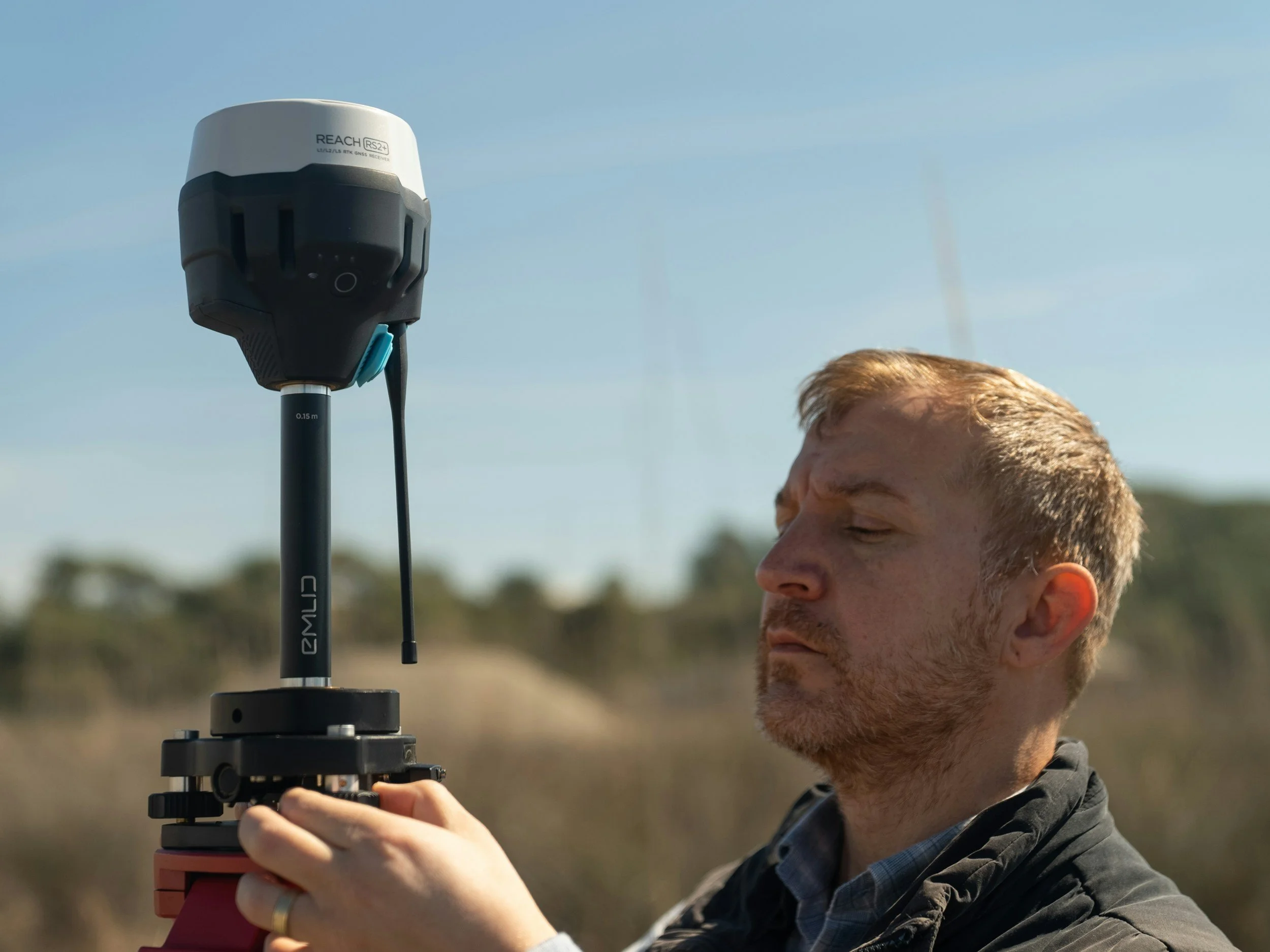 A man operating a surveying instrument outdoors during daytime with a clear sky and blurred natural background.