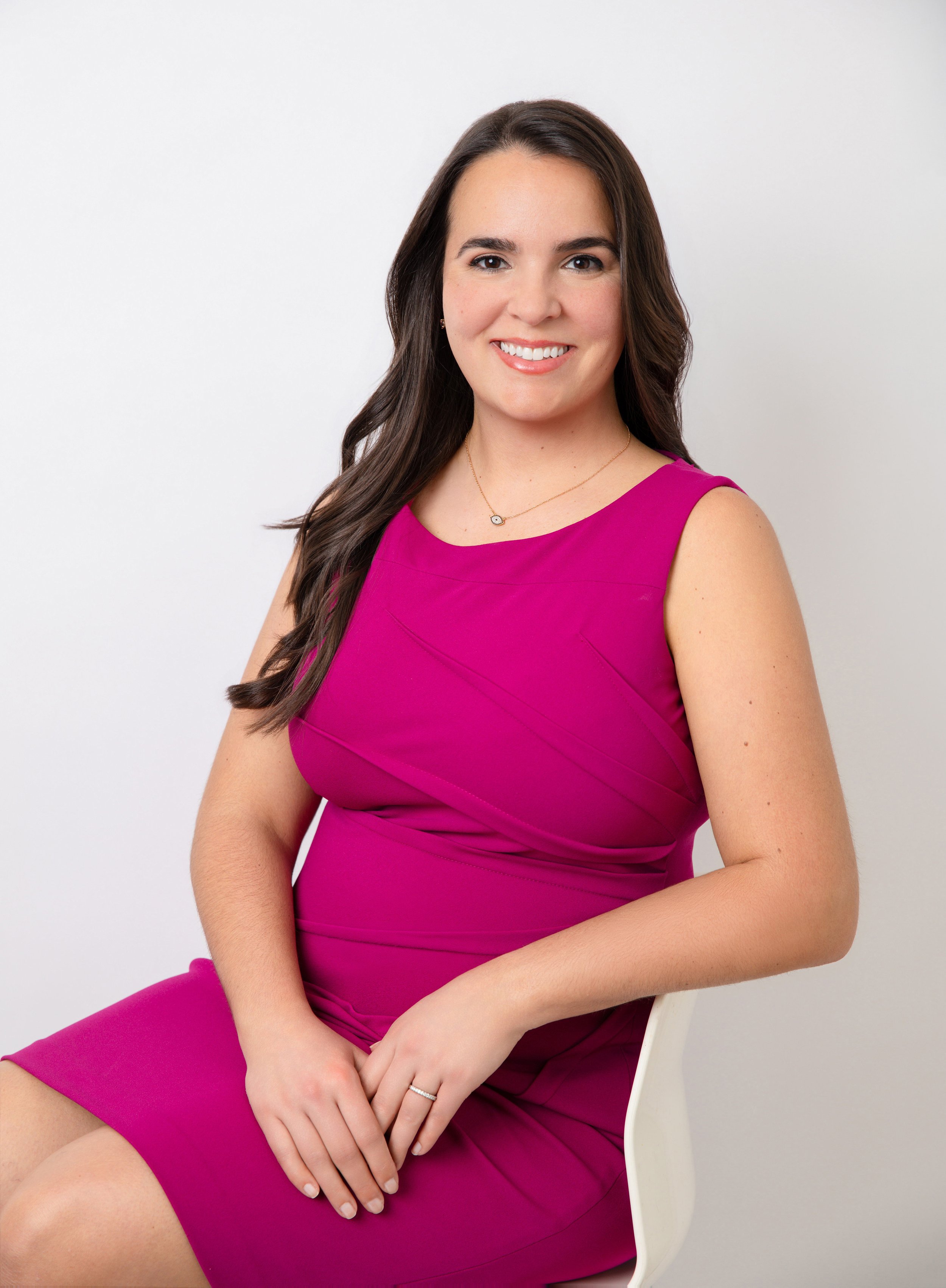A woman with long brown hair, wearing a bright pink dress and a necklace with a small pendant, is sitting on a white chair against a plain white background. She is smiling and looking at the camera.