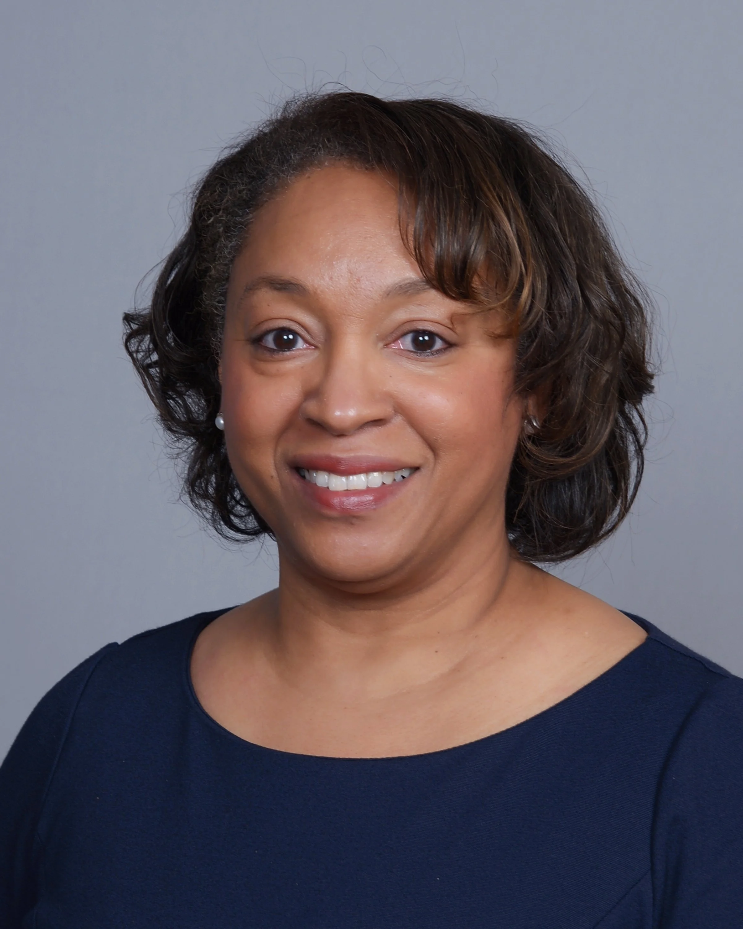 Headshot of a smiling woman with short, wavy hair, wearing pearl earrings and a navy blue top, against a plain gray background.