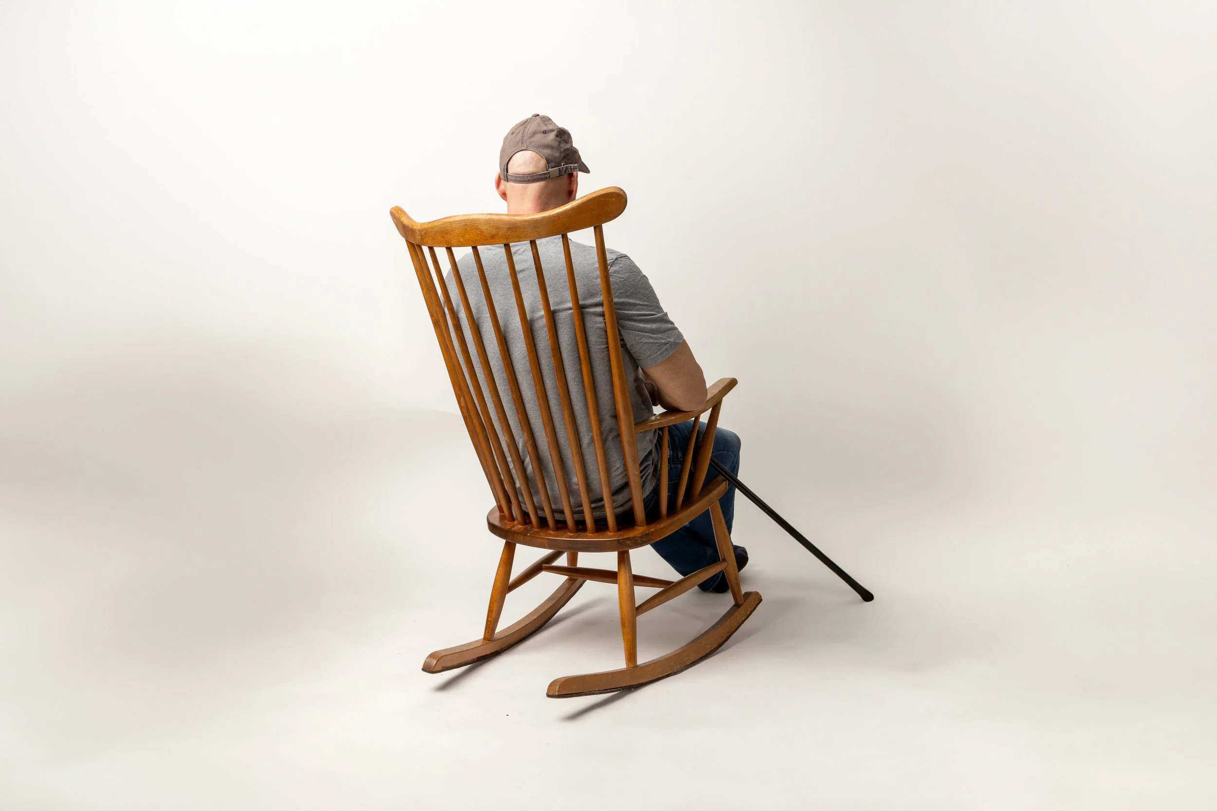 A man sitting in a wooden rocking chair with a gray cap and gray T-shirt, facing away from camera, in a plain white studio background.
