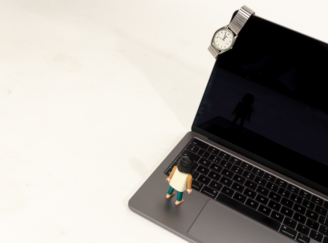 A laptop with a small figurine standing on the keyboard, a watch resting on the top edge of the monitor, and a silhouette of a person reflected on the screen.