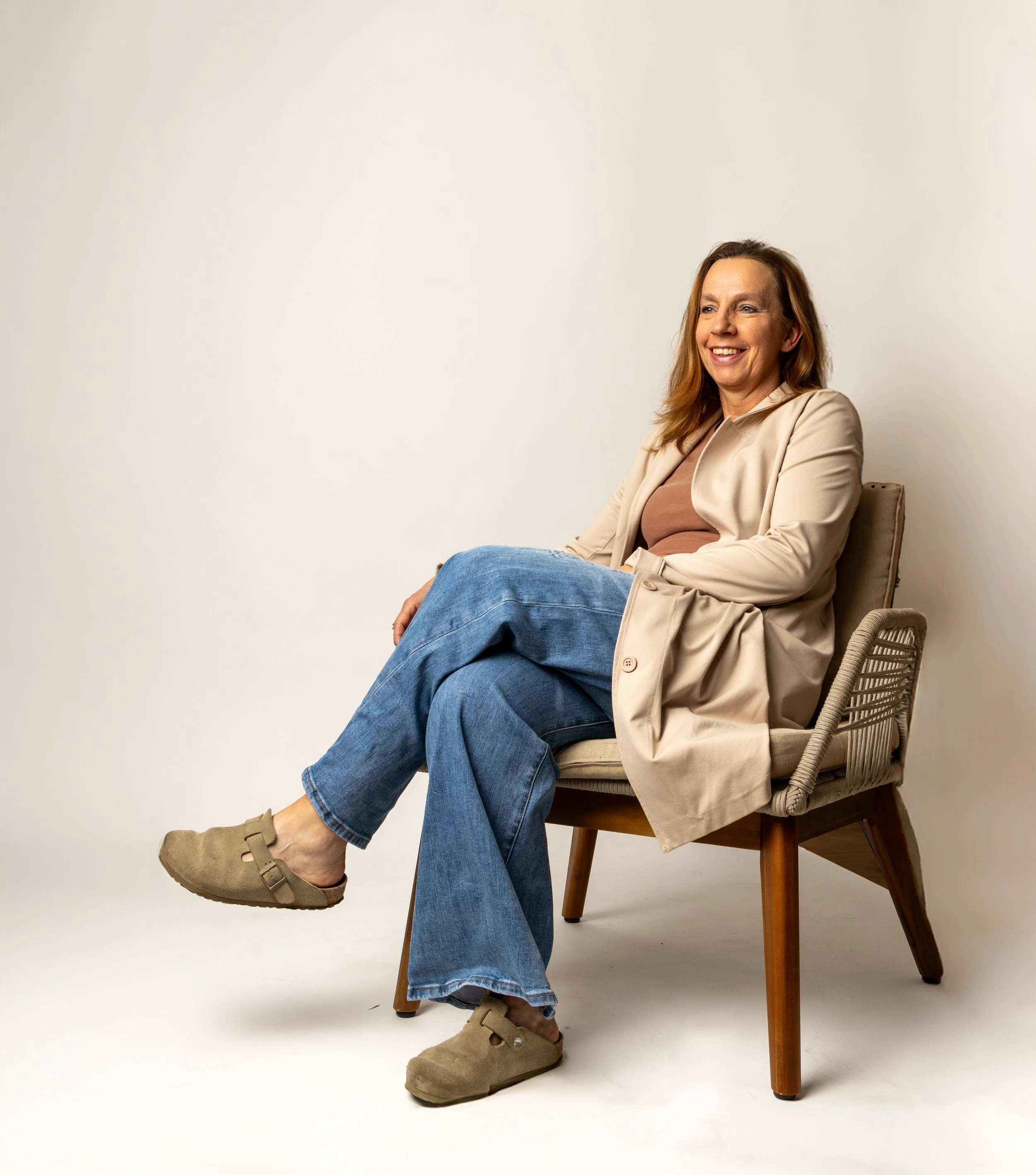 A middle-aged woman sitting on a beige chair, smiling, wearing a beige coat, brown top, blue jeans, and slippers, against a plain beige background.