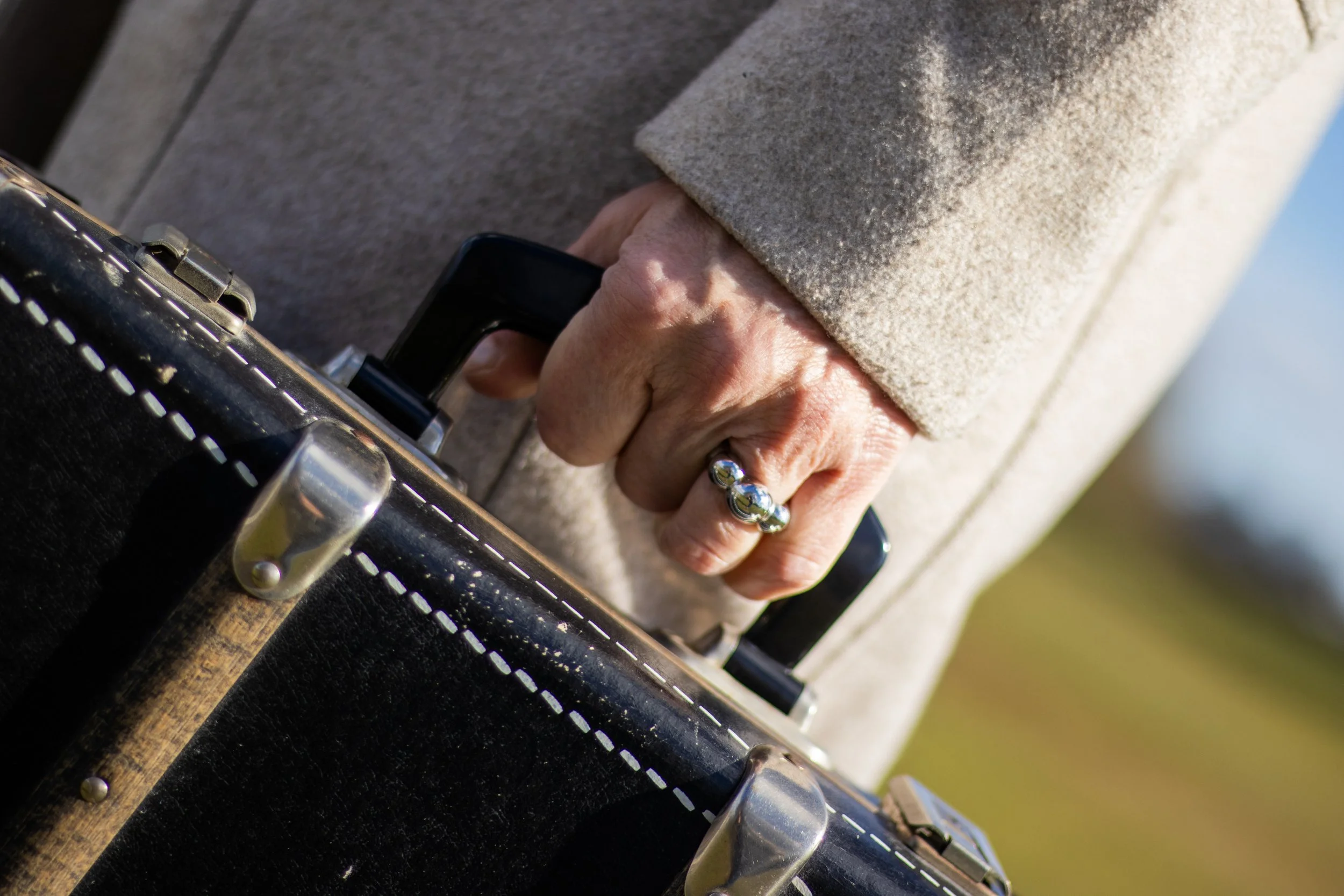 Close-up of a person's hand holding a black vintage suitcase, wearing a silver ring with multiple beads, and dressed in a beige coat, against a blurred background.