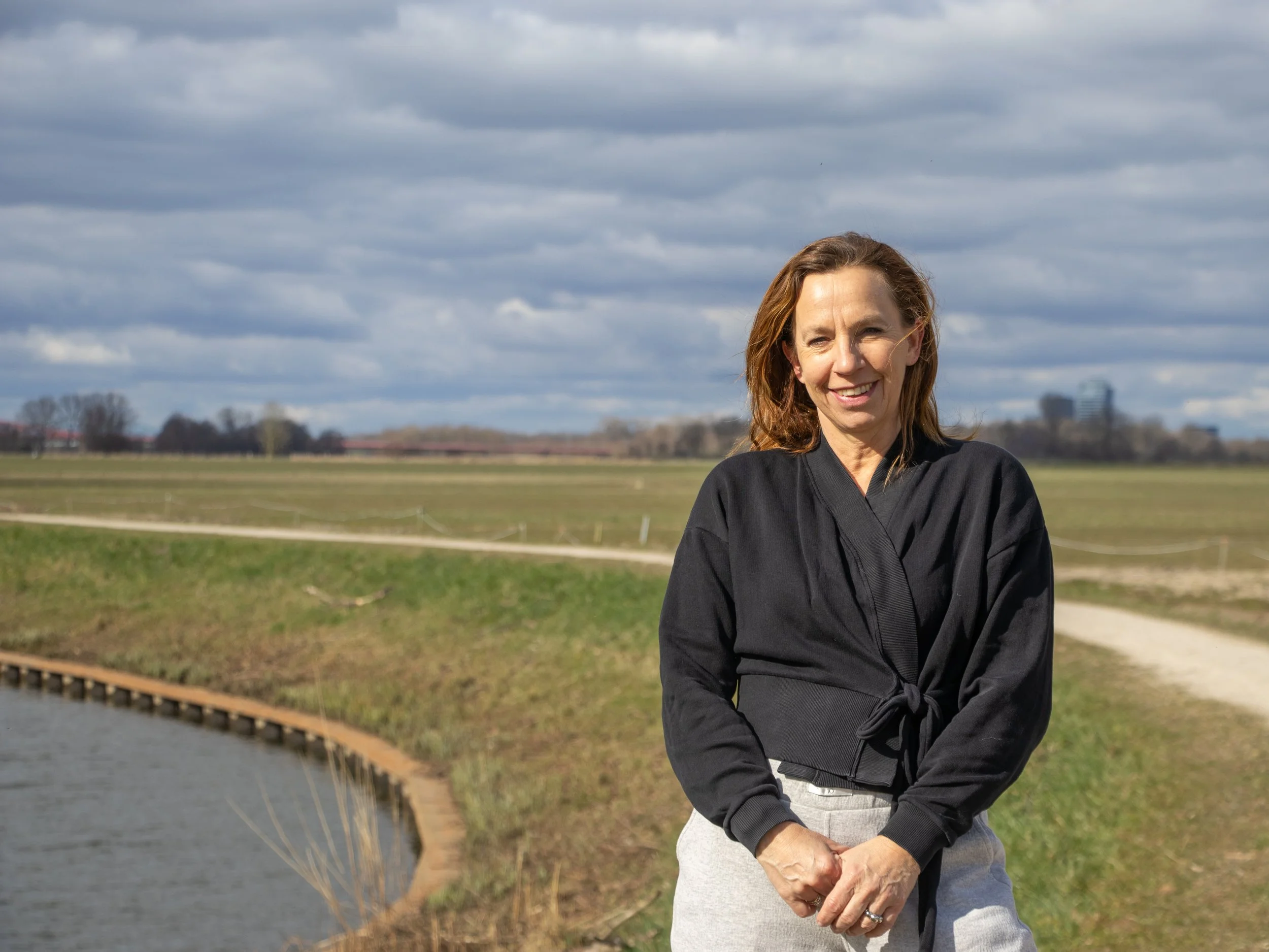 A woman smiling outdoors near a small pond, with grassy fields and a cloudy sky in the background.