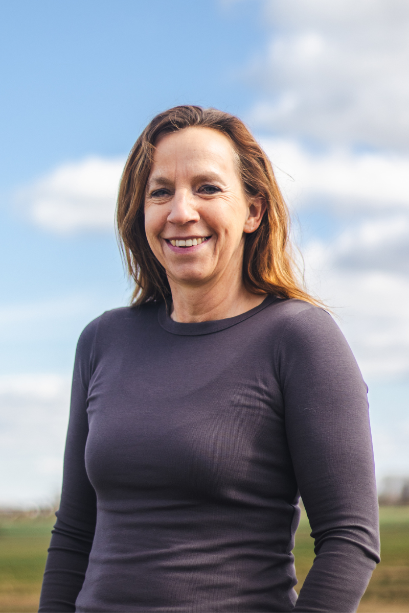 Portrait of a woman with shoulder-length brown hair smiling outdoors on a partly cloudy day.