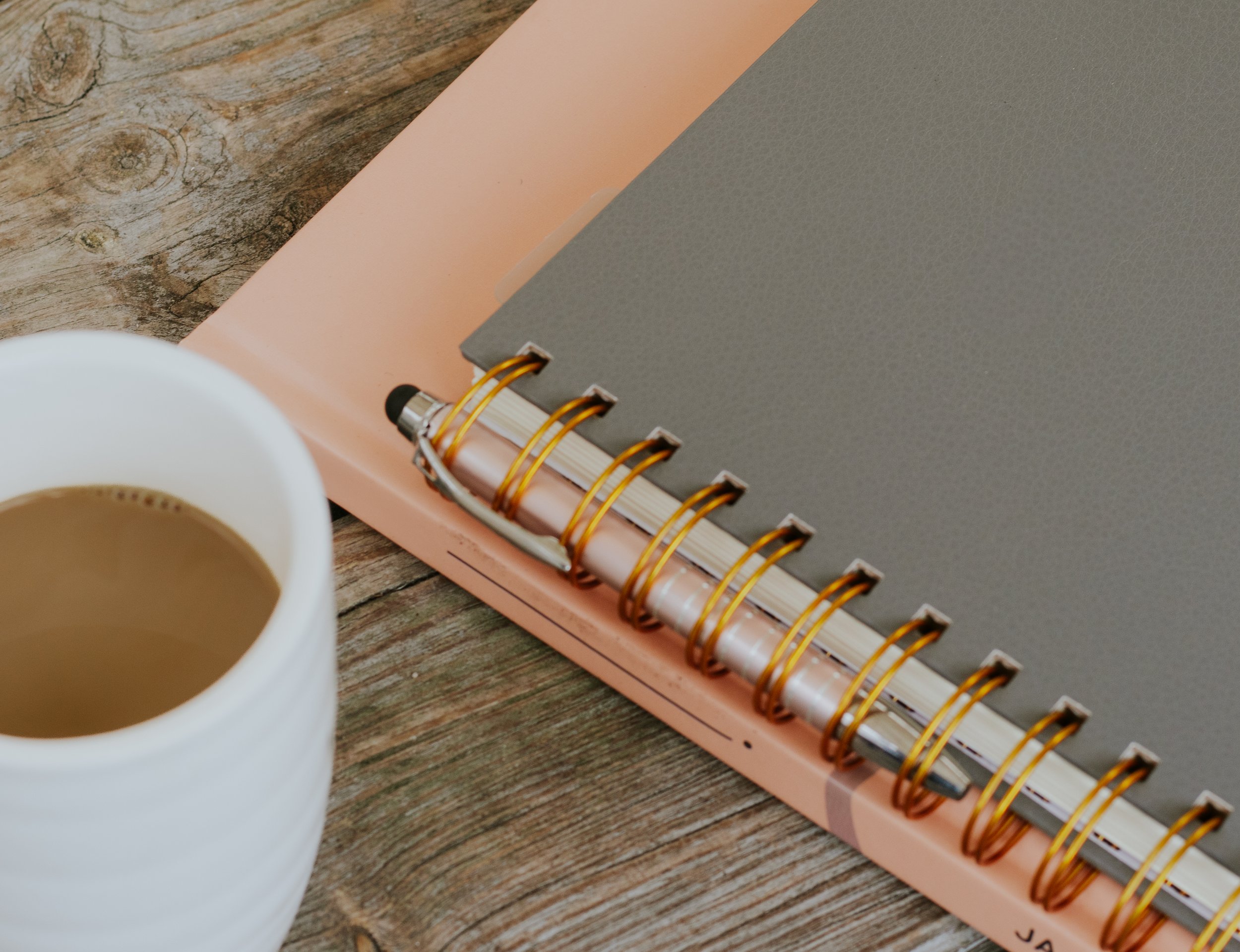 A close-up of a cup of coffee on a wooden table next to a pink and gray spiral-bound notebook.
