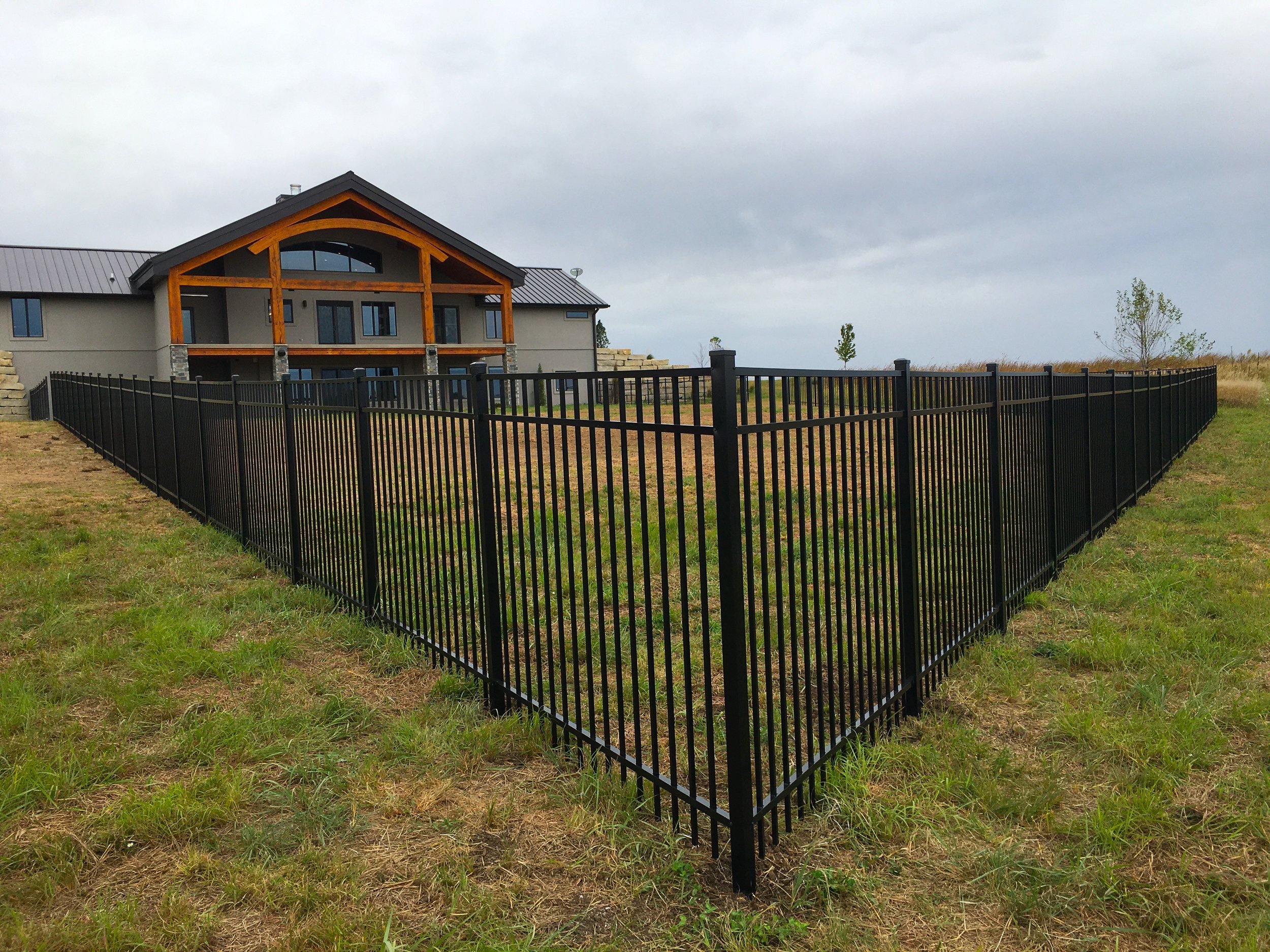Residential building with triangle roof and large windows, surrounded by black metal fence, on grassy yard under cloudy sky.