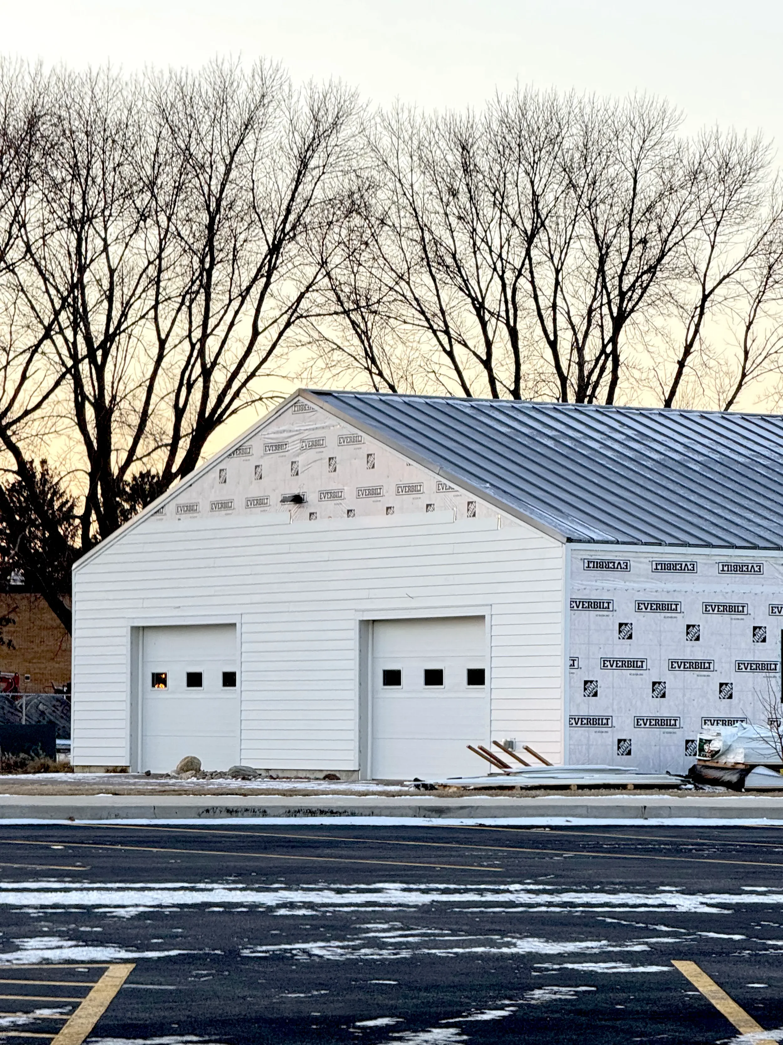 Under-construction garage with white siding and metal roof, surrounded by leafless trees and a parking lot with patches of snow.