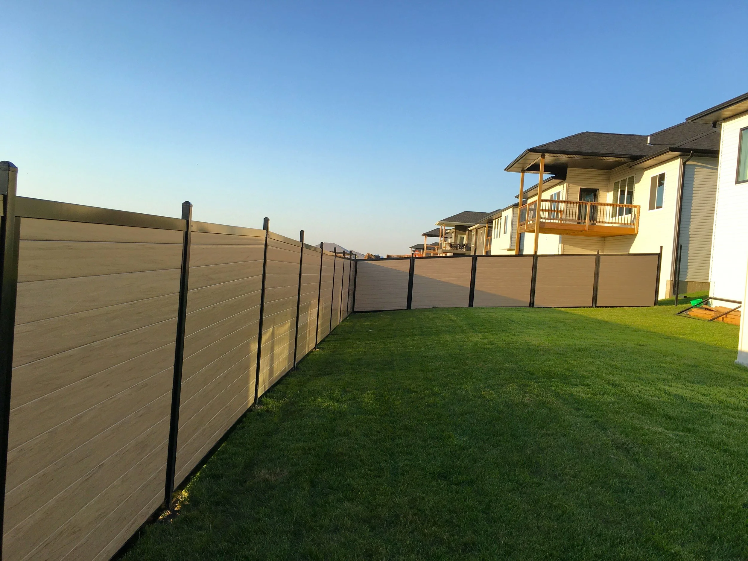 Residential backyard with a wooden privacy fence and grass lawn, next to modern houses with balconies under a clear blue sky.