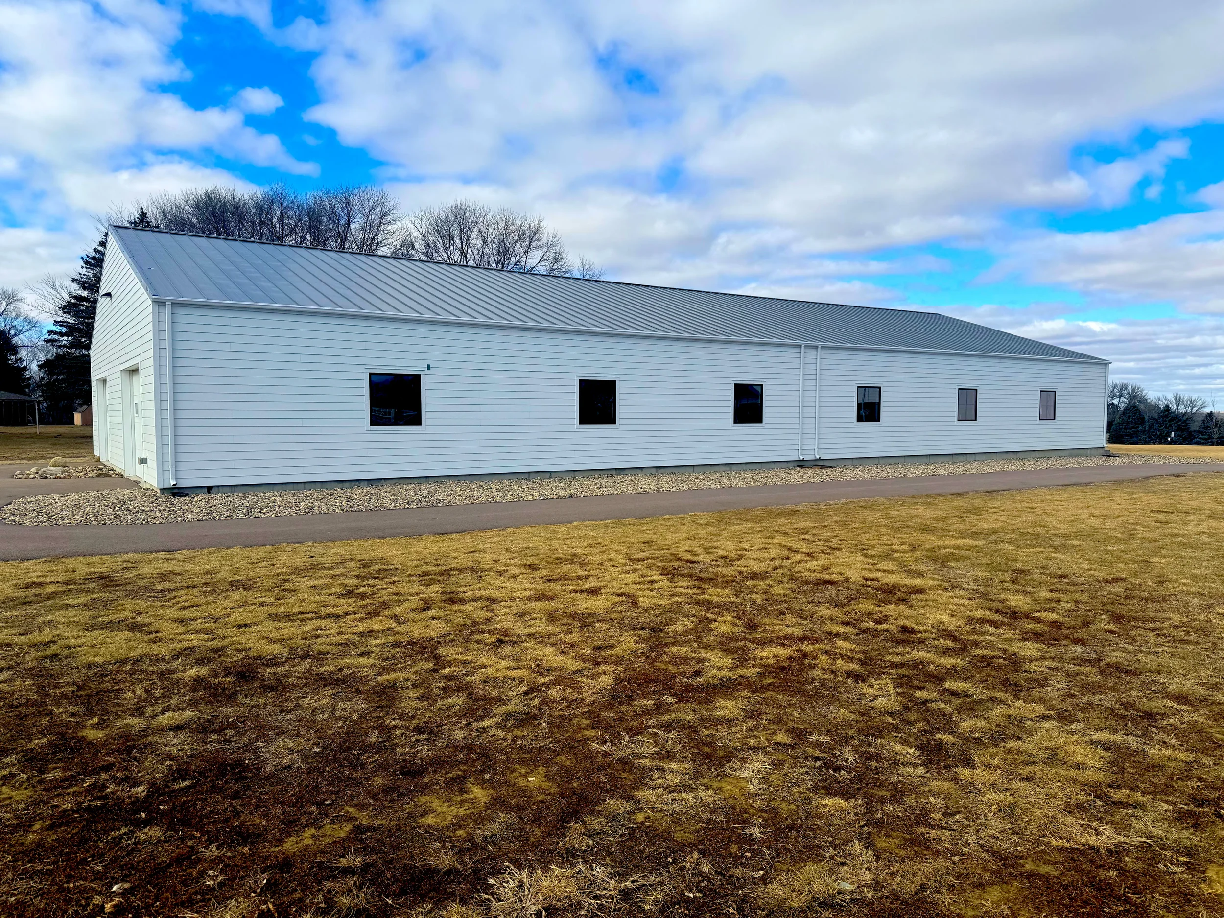 Exterior of a long, white metal building with a pitched roof, situated in a grassy field with a paved walkway in front, under a partly cloudy sky.
