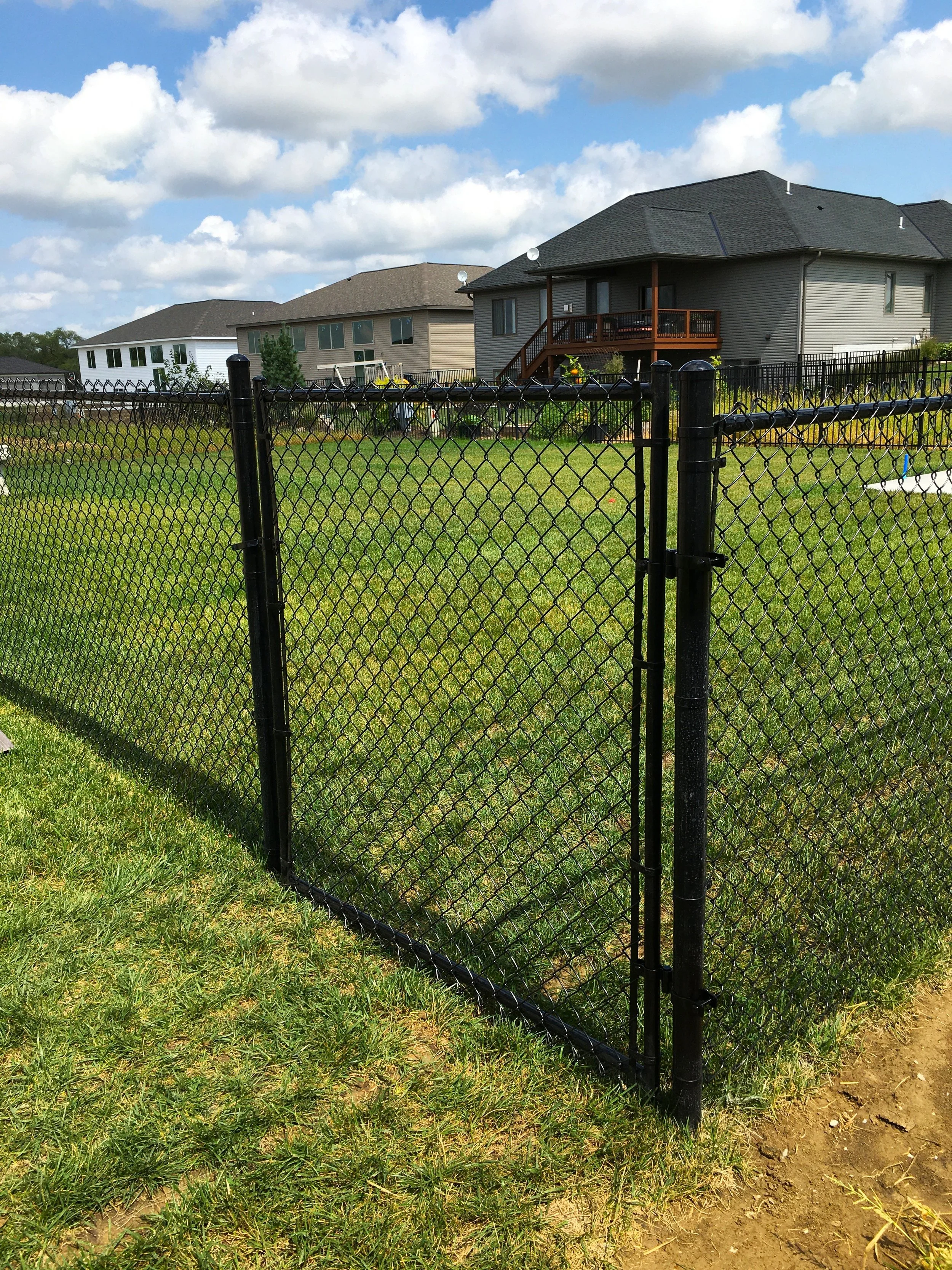 Black chain-link fence with gate in a grassy yard, suburban houses in the background, partly cloudy sky overhead.