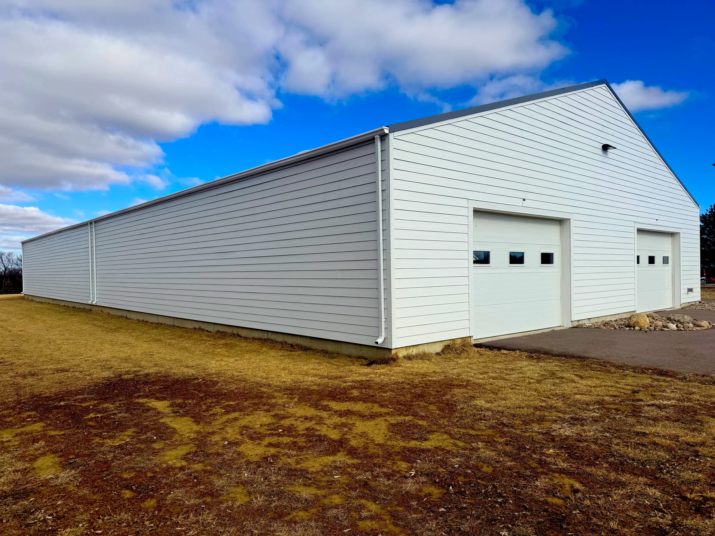A large white metal building with a gable roof and two garage doors, set on a grassy lot with a paved area in front, under a partly cloudy sky.