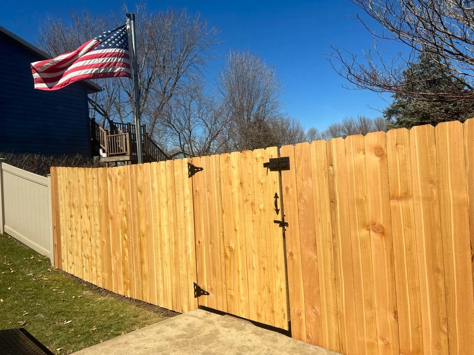 Wooden fence with a latch in a backyard, featuring an American flag on a pole in the background near a house with visible trees under a clear blue sky.