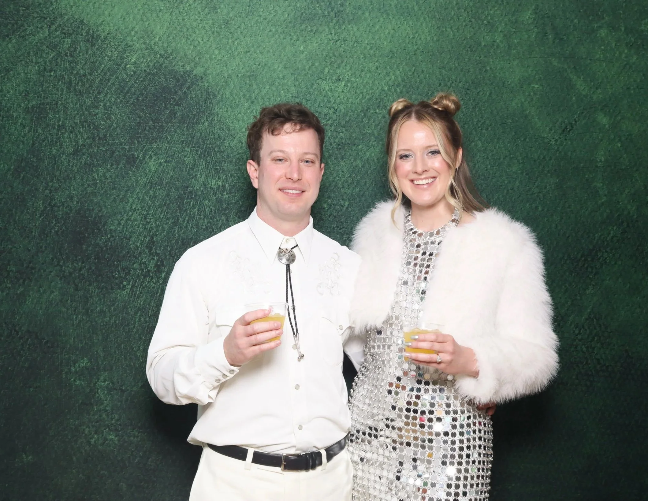 A man and woman dressed in formal attire holding drinks and smiling at a photo event with a green textured backdrop behind them.