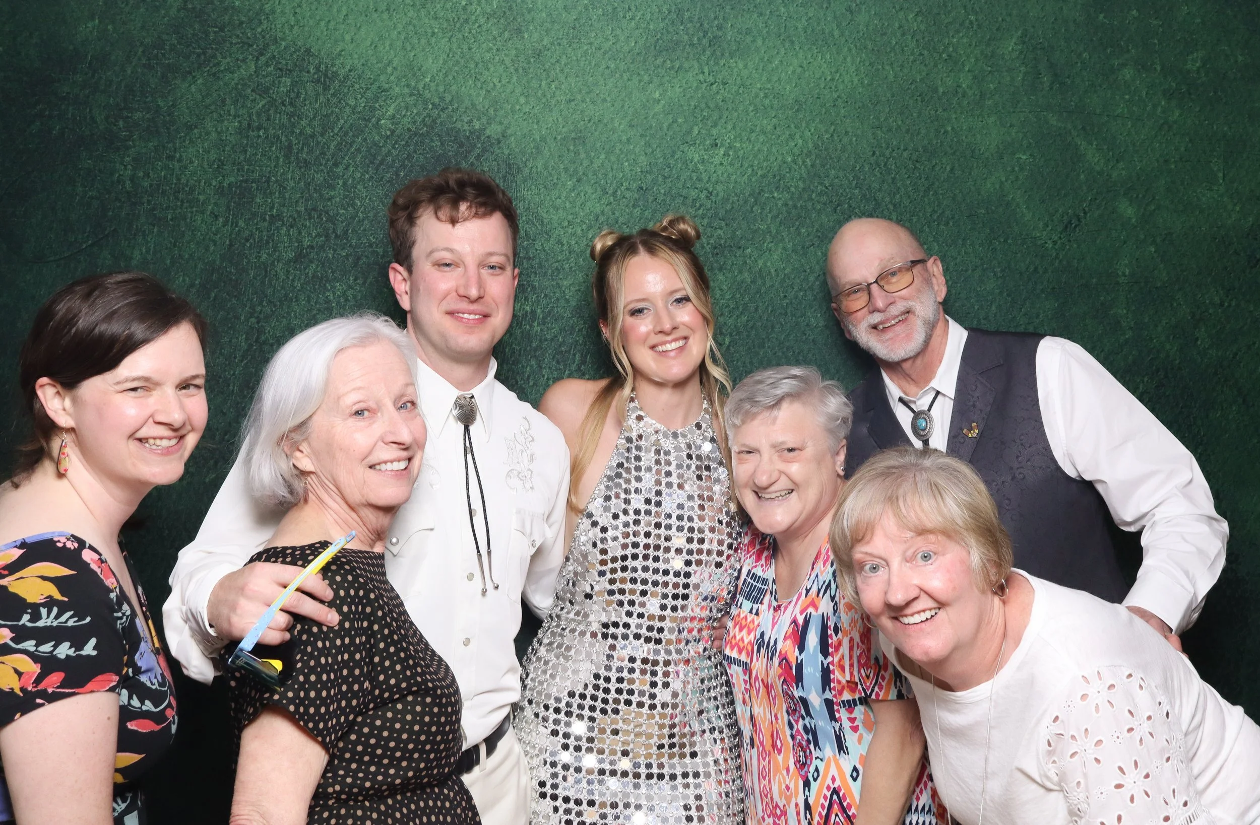 A group of people smiling together in front of a green textured backdrop, some wearing colorful clothing and accessories.