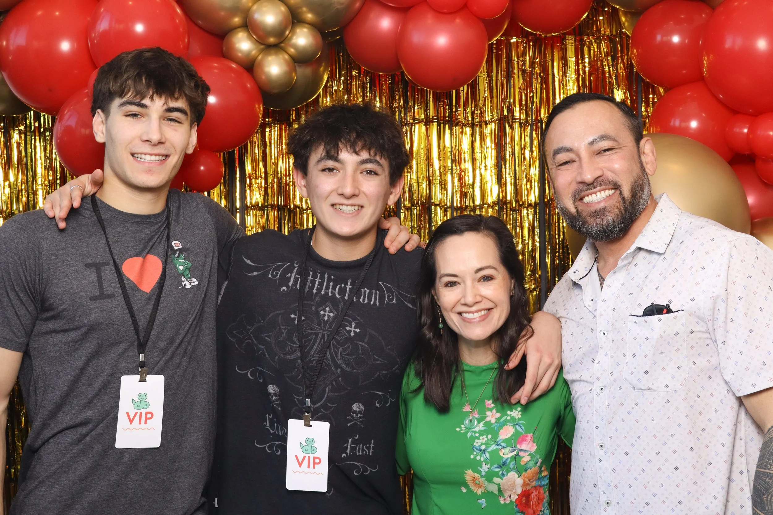 Group photo of four people in front of a red and gold balloon backdrop with VIP tags.