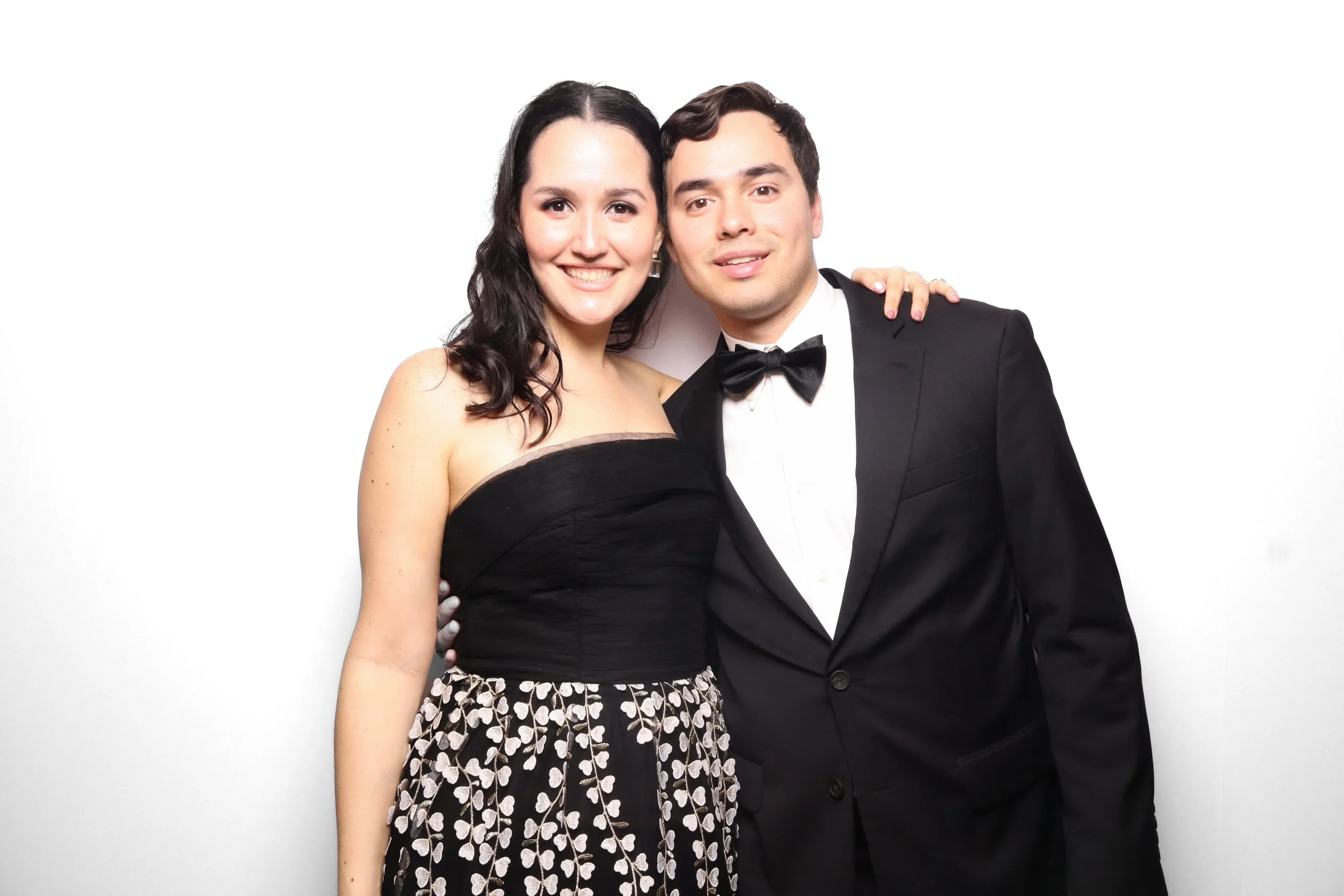 A smiling woman in a black dress with a floral skirt and a man in a tuxedo with a bow tie, standing closely together against a white background.