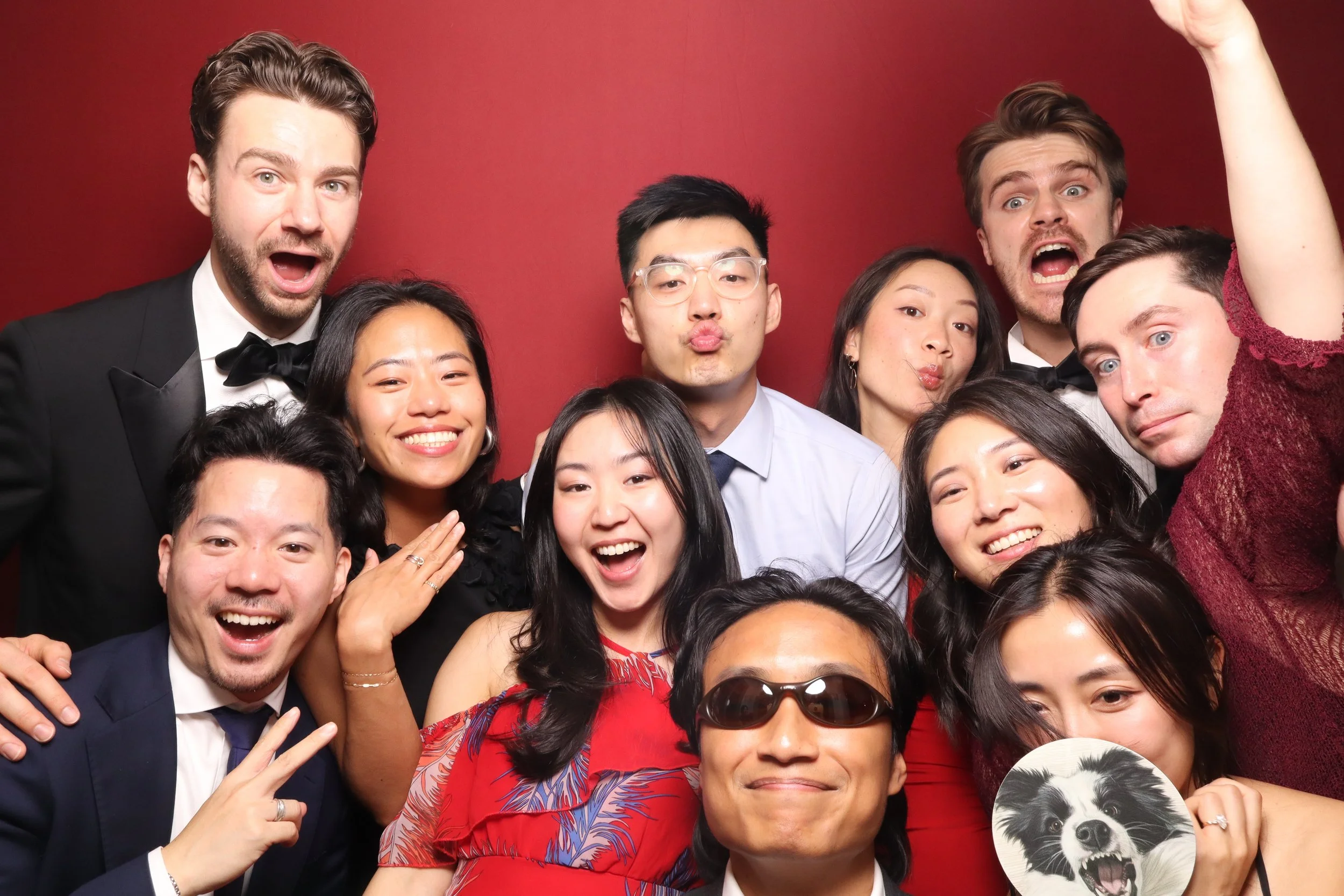 A group of diverse people posing for a photo at a celebration or event against a red background, displaying happy and excited expressions.