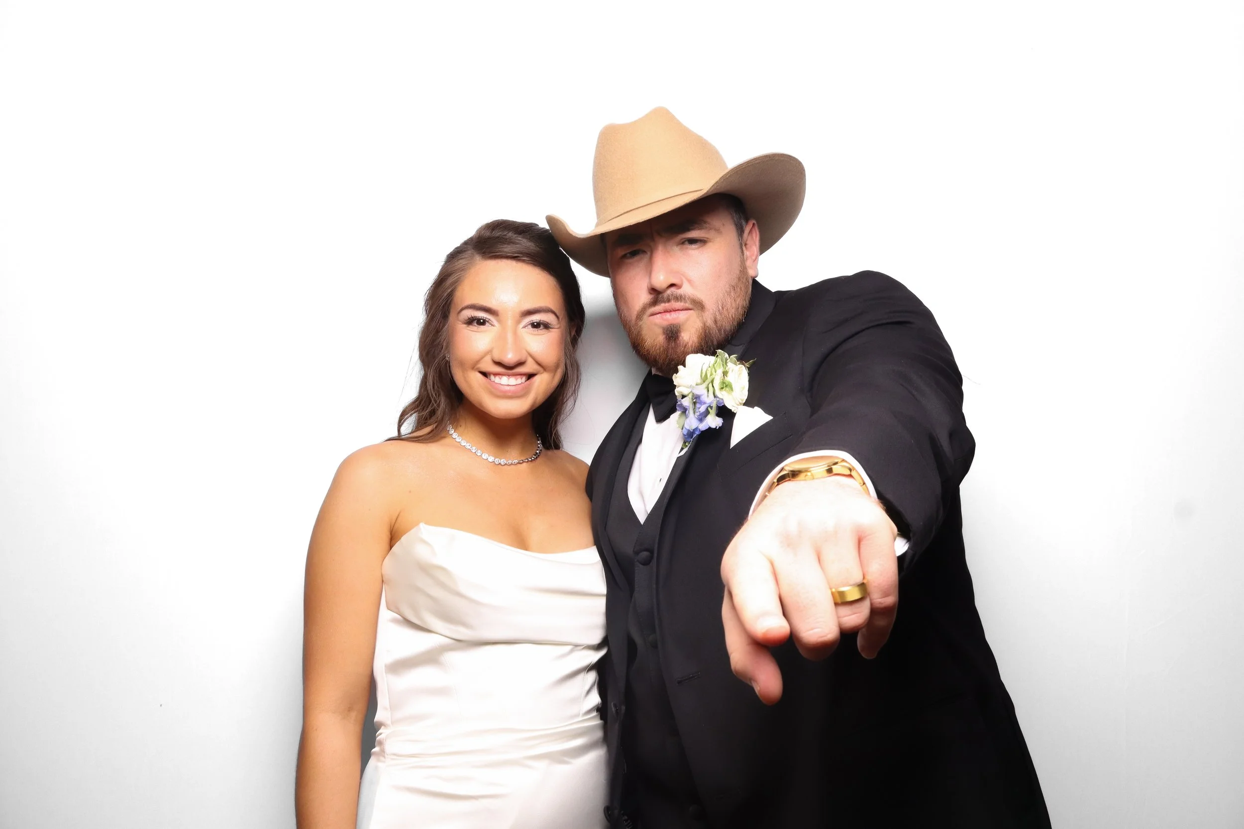 A man and woman dressed in wedding attire, taking a selfie against a plain white background. The woman is wearing a strapless white wedding dress and a pearl necklace, smiling, while the man is wearing a black tuxedo with a boutonnière and a cowboy h