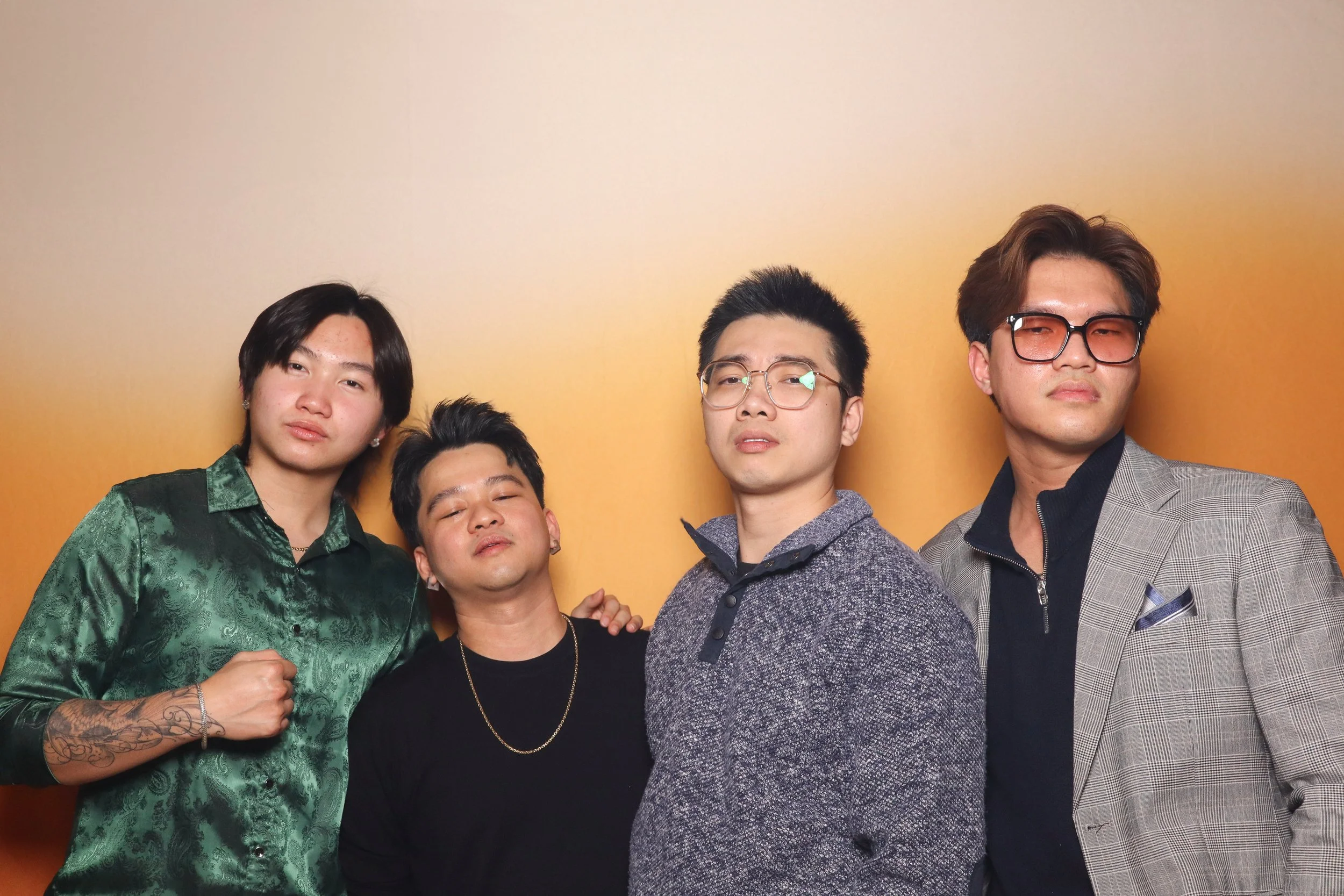 Four men posing in front of a gradient background; one is wearing a green shirt, another in a black shirt, one in a grey sweater, and the last in a grey jacket with sunglasses.