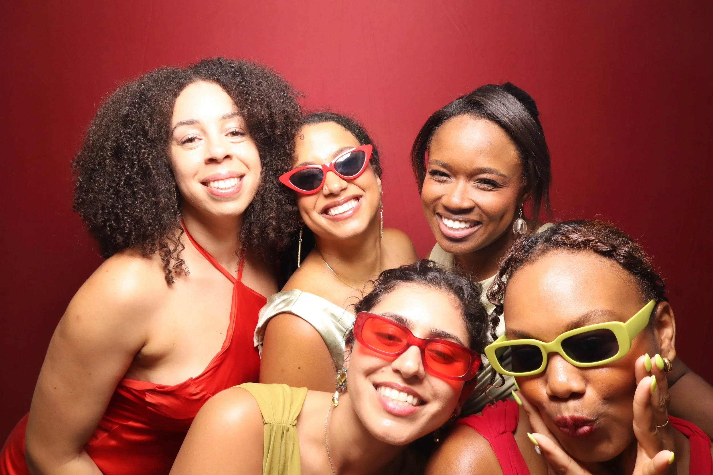 Group of women smiling, wearing colorful clothing and sunglasses, standing in front of a red background.