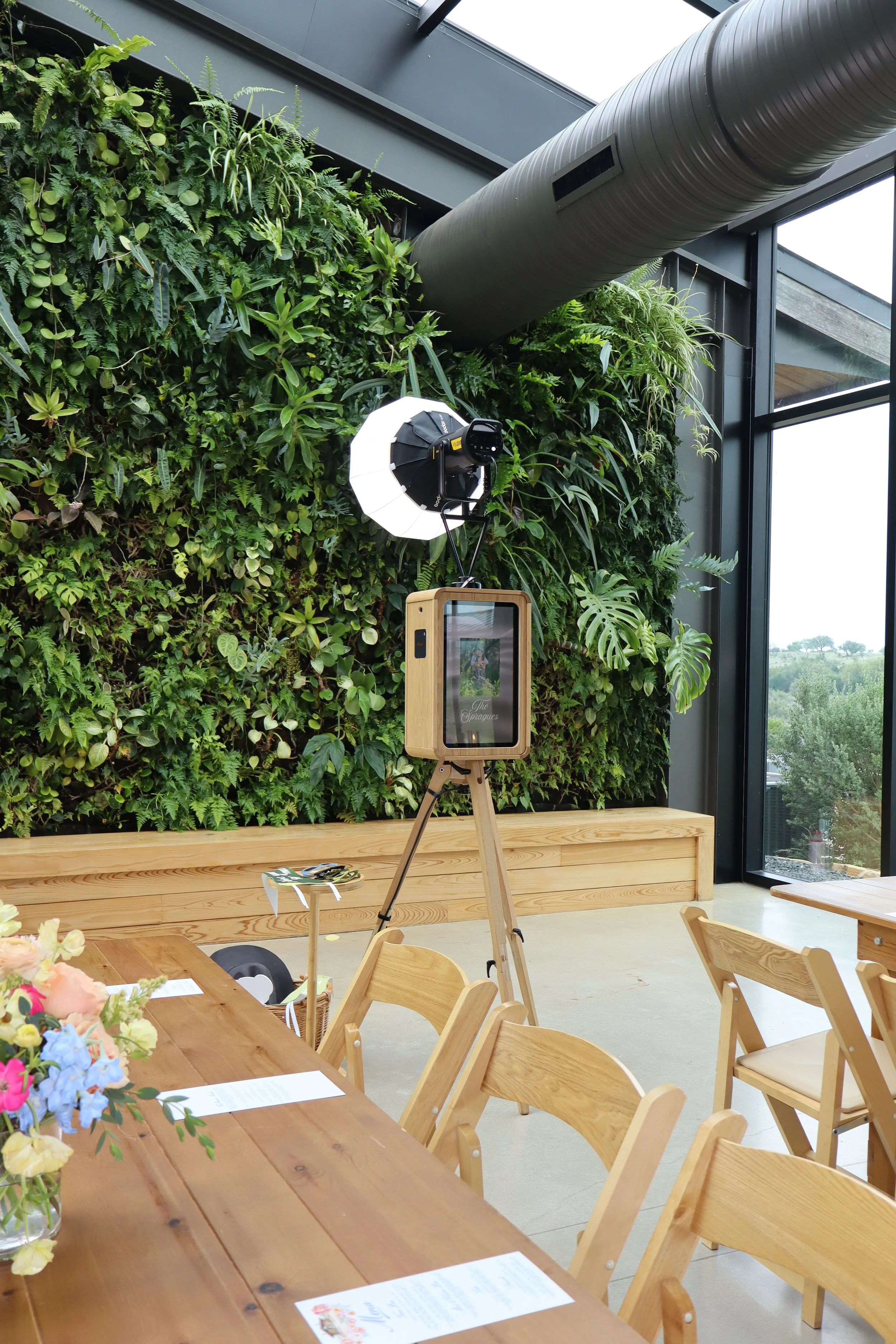 Interior of a modern space with a green living wall, wooden tables and chairs, a photo booth setup, large windows showing a view of trees, and a ventilation duct on the ceiling.