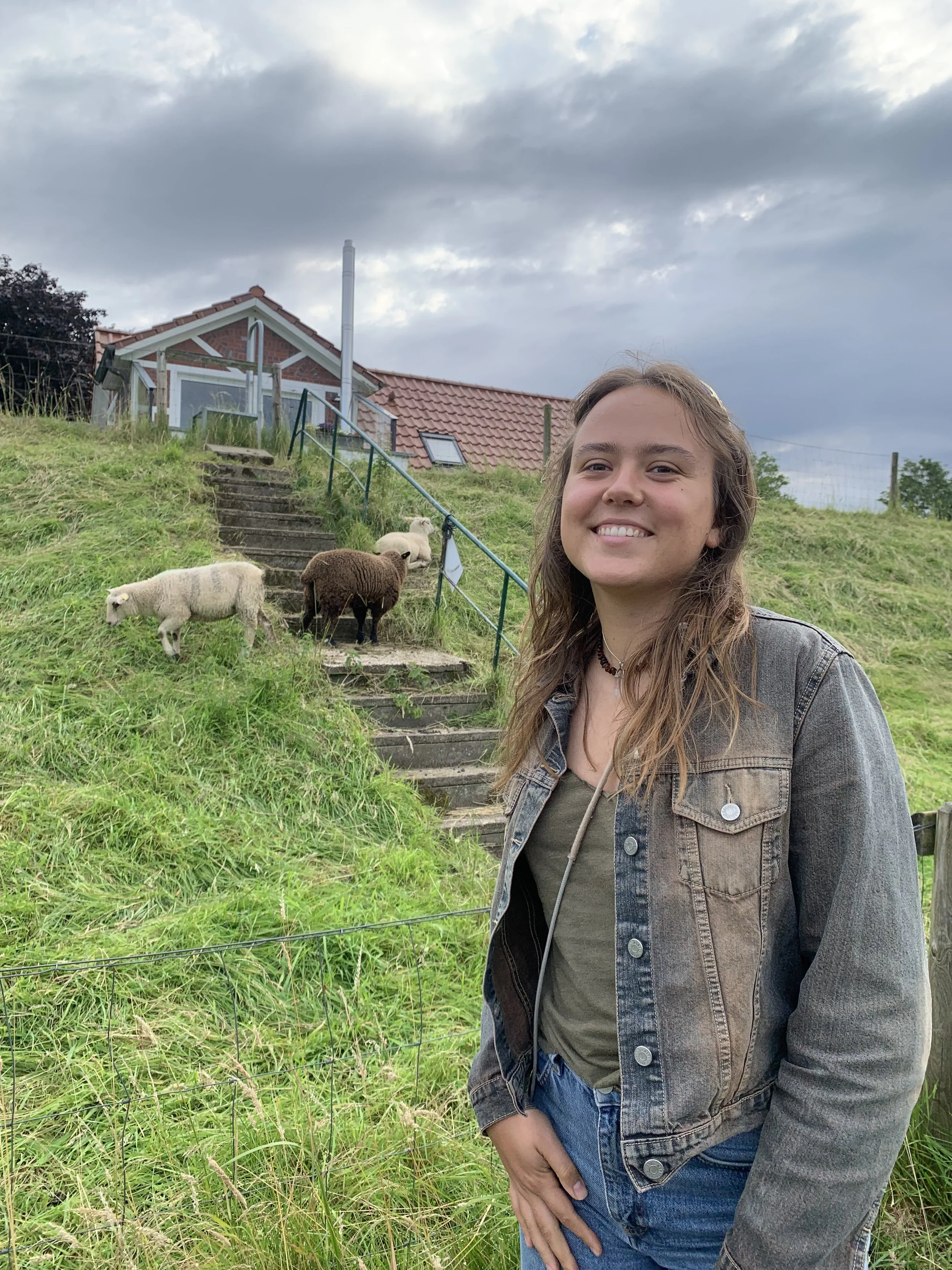 Eine lachende junge Frau steht vor einer Weide mit Schafen, im Hintergrund ein Landhaus bei bewölktem Himmel.