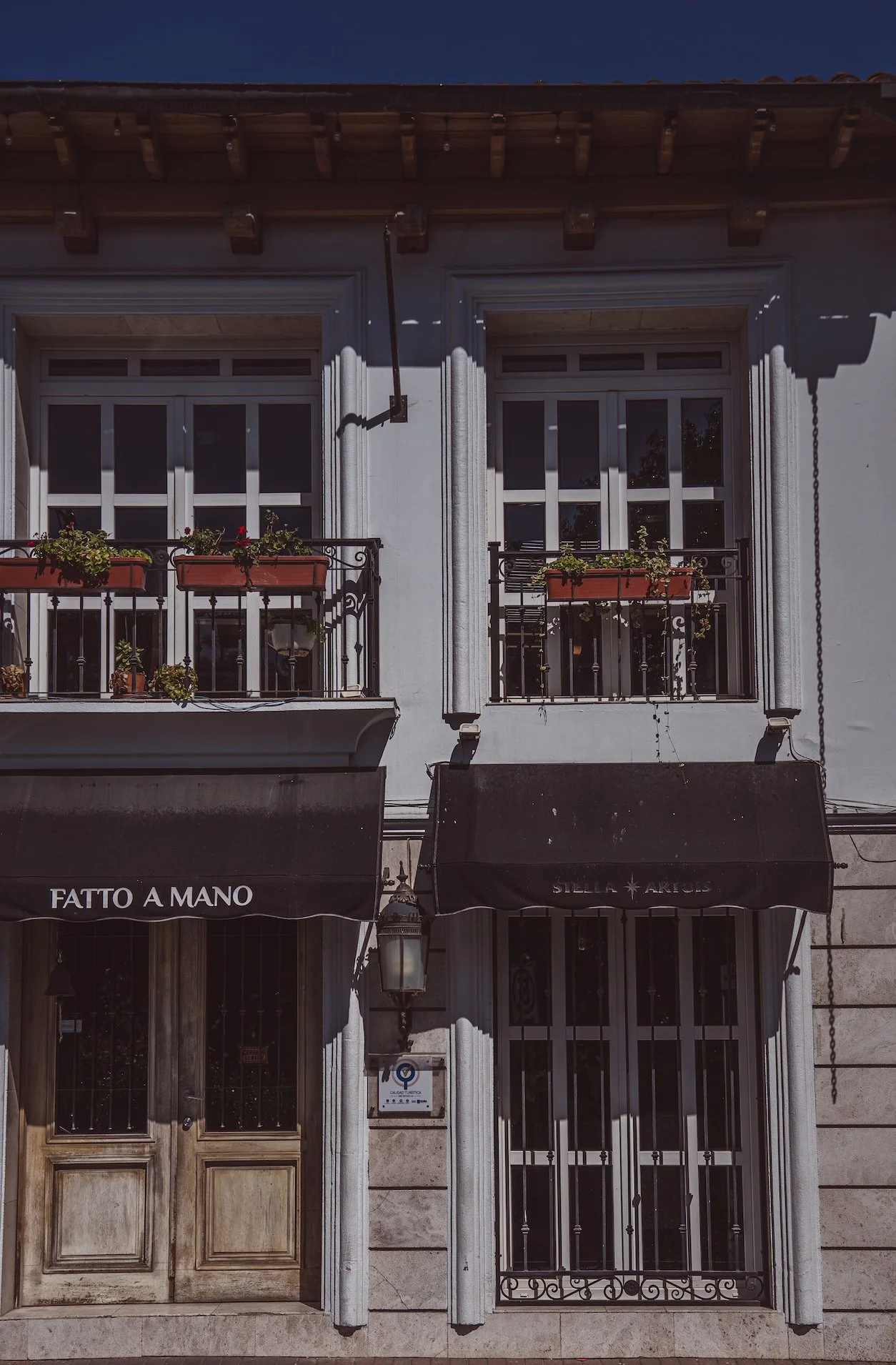 Edificio de dos pisos con balcones con macetas de flores y toldos negros en la fachada principal, puertas de madera y rejas de hierro.