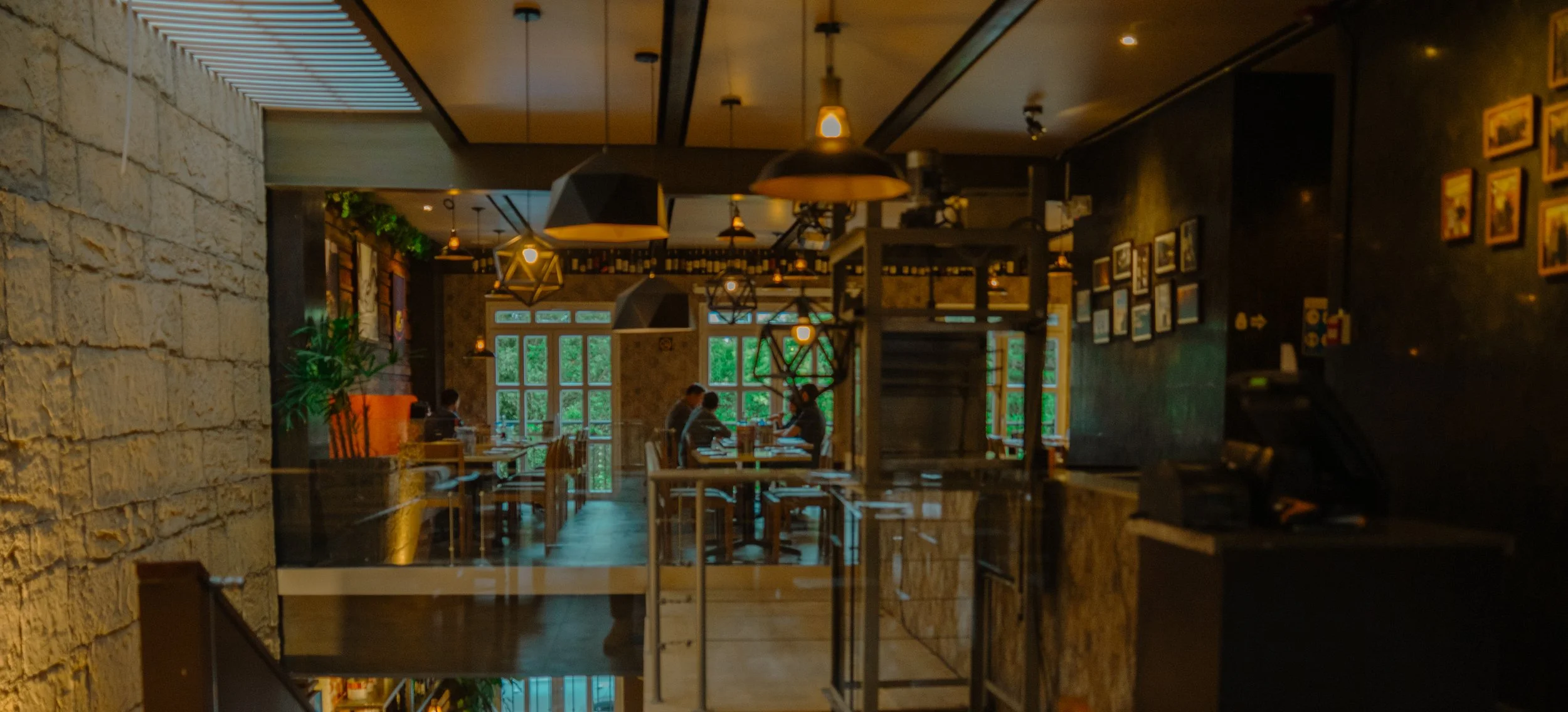 Interiores de un restaurante acogedor con paredes de piedra y decoración con cuadros en la pared, mesas y sillas de madera, y varias personas conversando en un ambiente cálido y bien iluminado con lámparas colgantes.